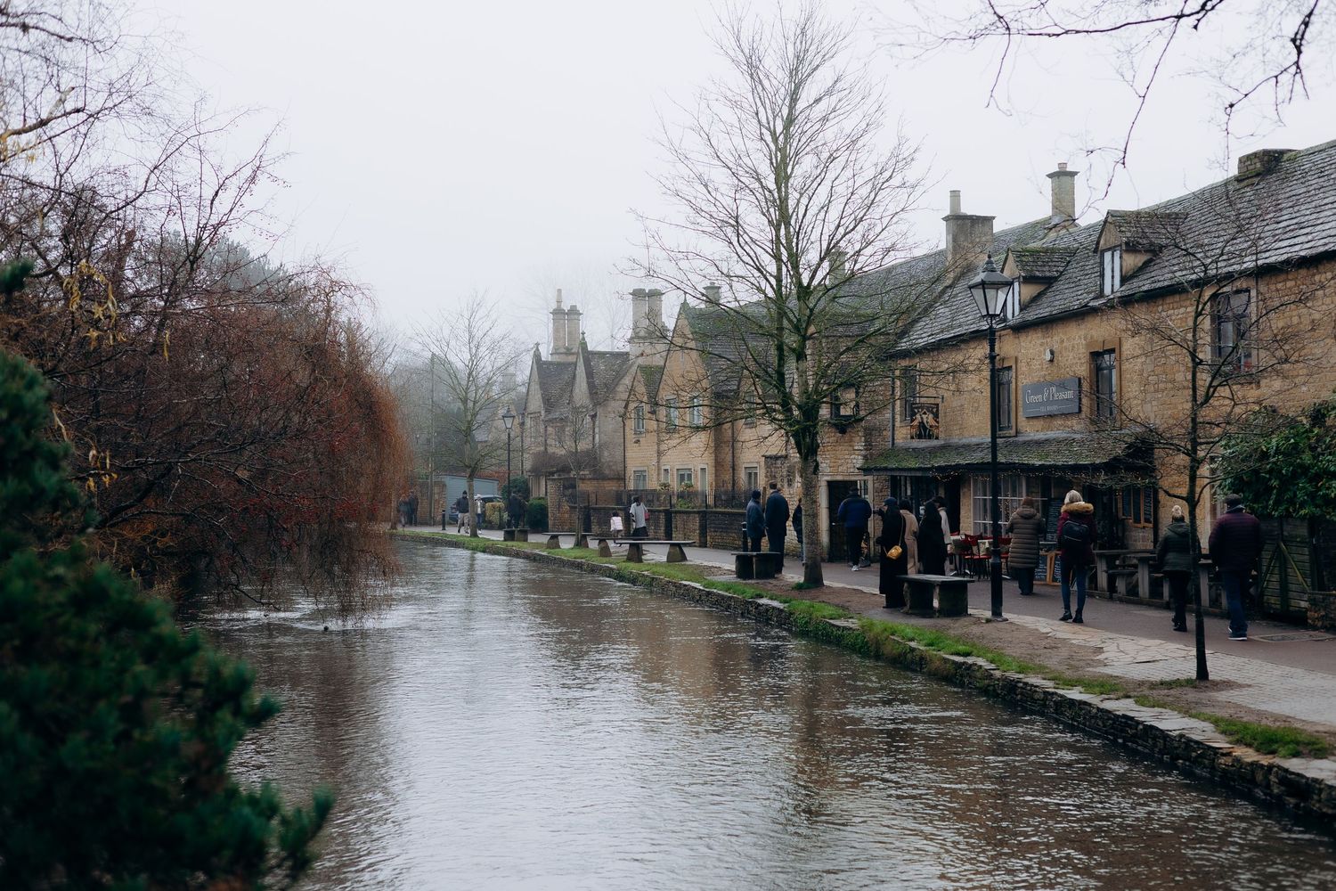 People walking along a historic stone street with traditional Cotswold cottages on a cloudy day.