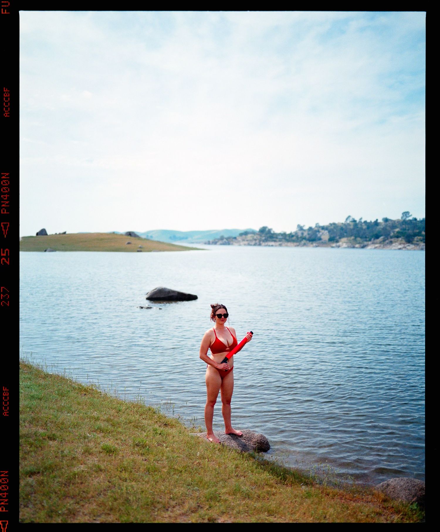 Woman in red bikini and cap standing by a lake with hills in the background on a film photograph.