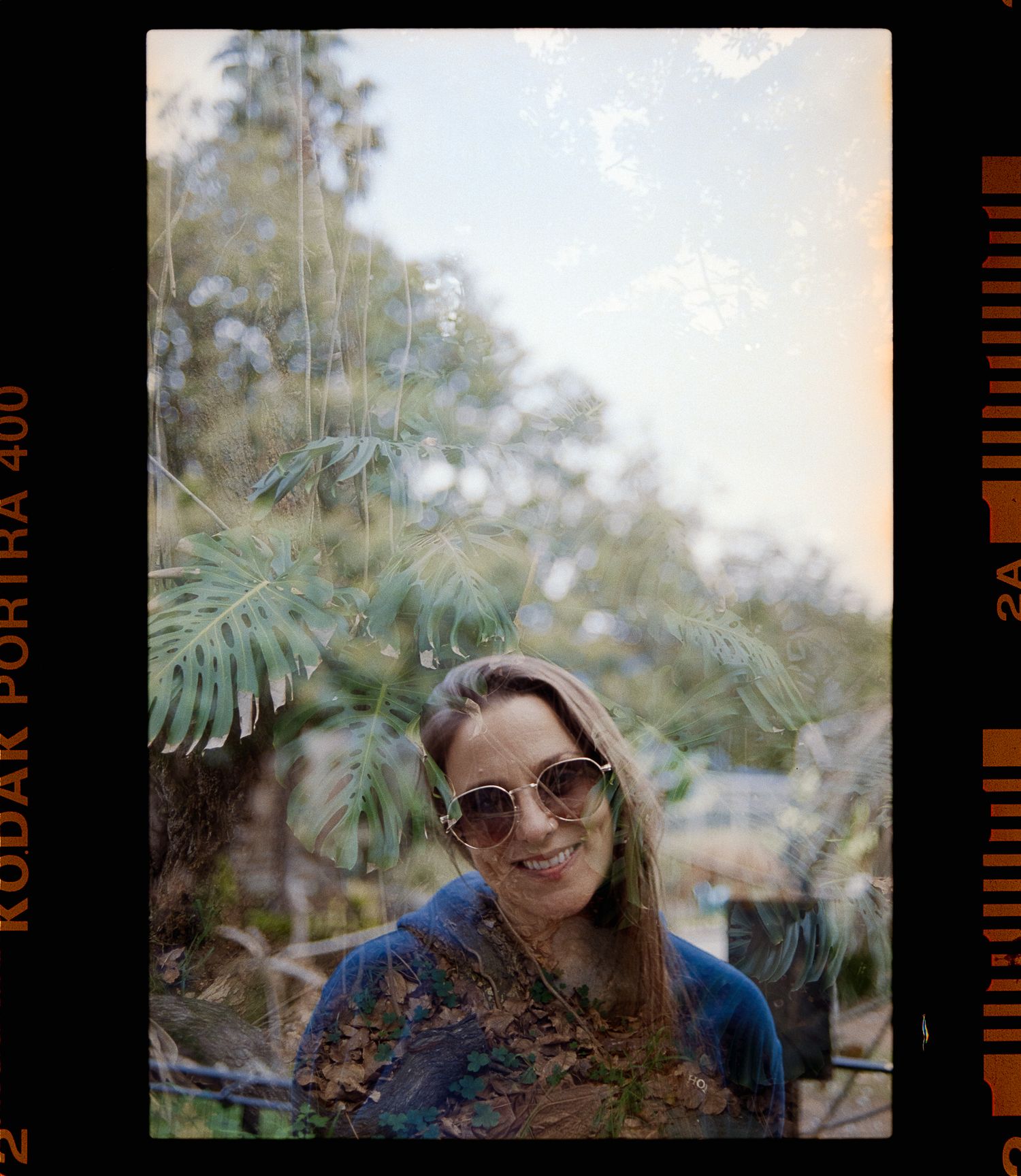 A person in sunglasses smiles while posing outdoors with blurred greenery and foliage in the background.