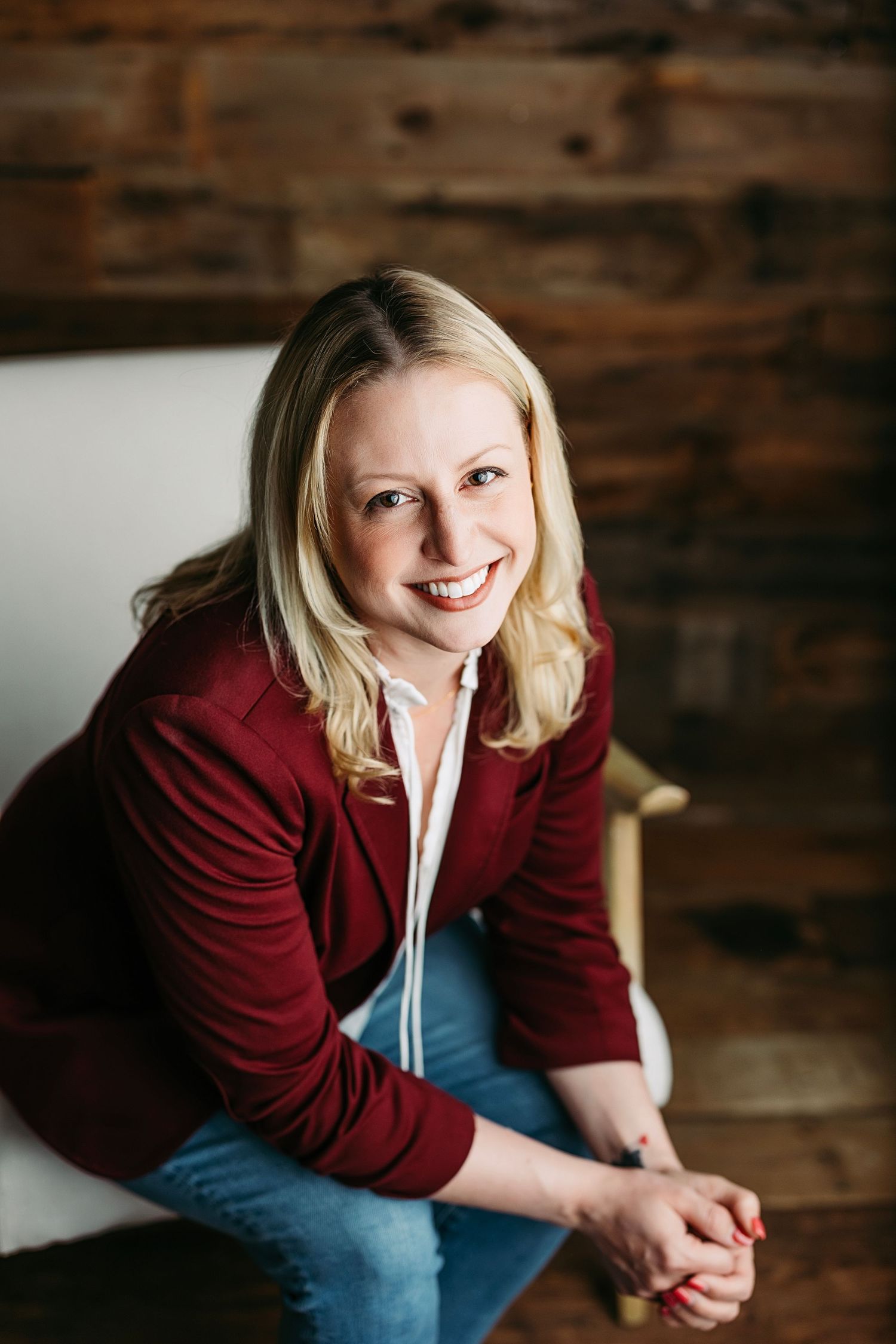 Smiling blonde woman in red blazer and jeans sitting in white chair against rustic wood wall background.