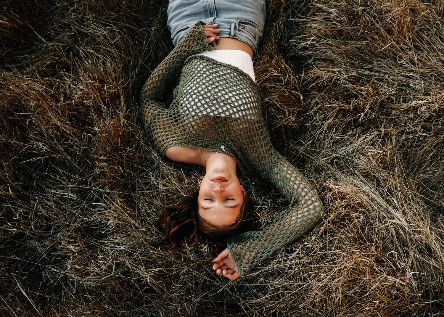 Woman lying on dry grass wearing fishnet tights and denim shorts, viewed from above in a serene outdoor setting.
