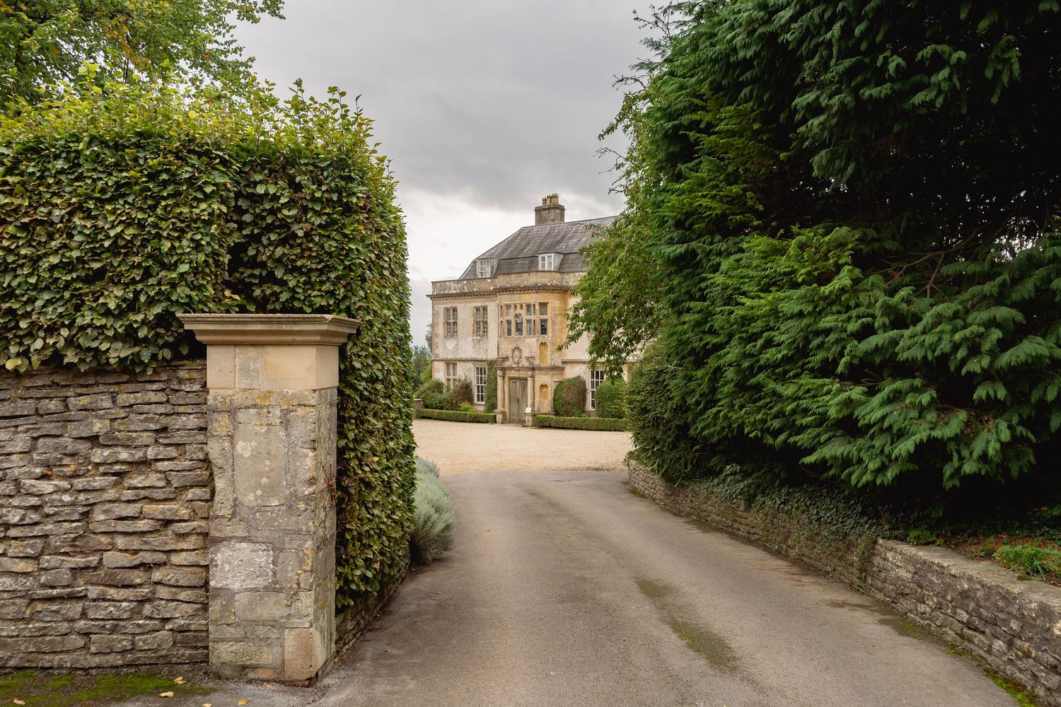 Stone gatepost and hedge-lined driveway leading to an elegant Georgian country manor house surrounded by mature trees. Hamswell House, Bath