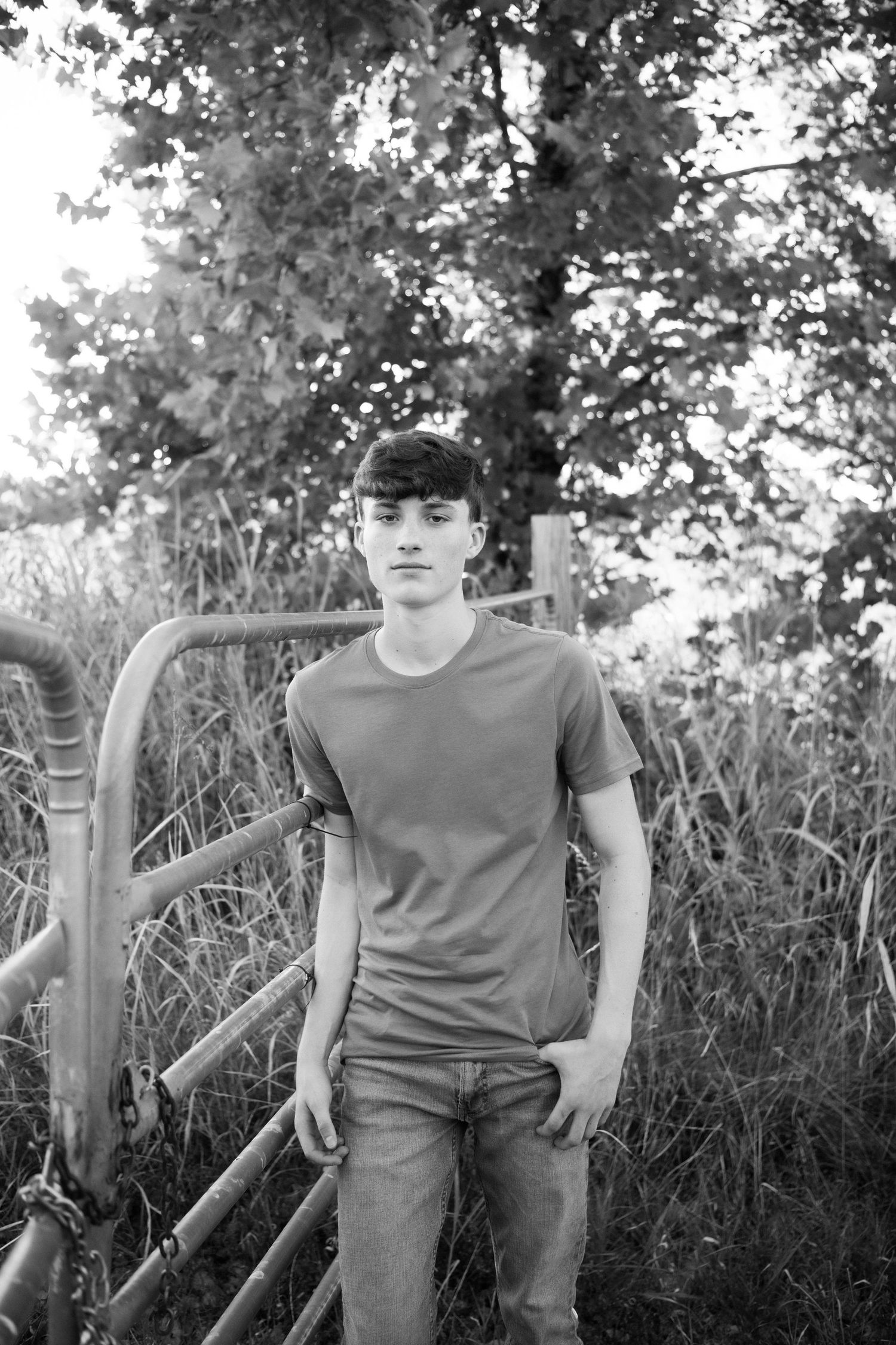 A person sits casually on a rustic wooden fence surrounded by tall grass and trees in a black and white photograph.
