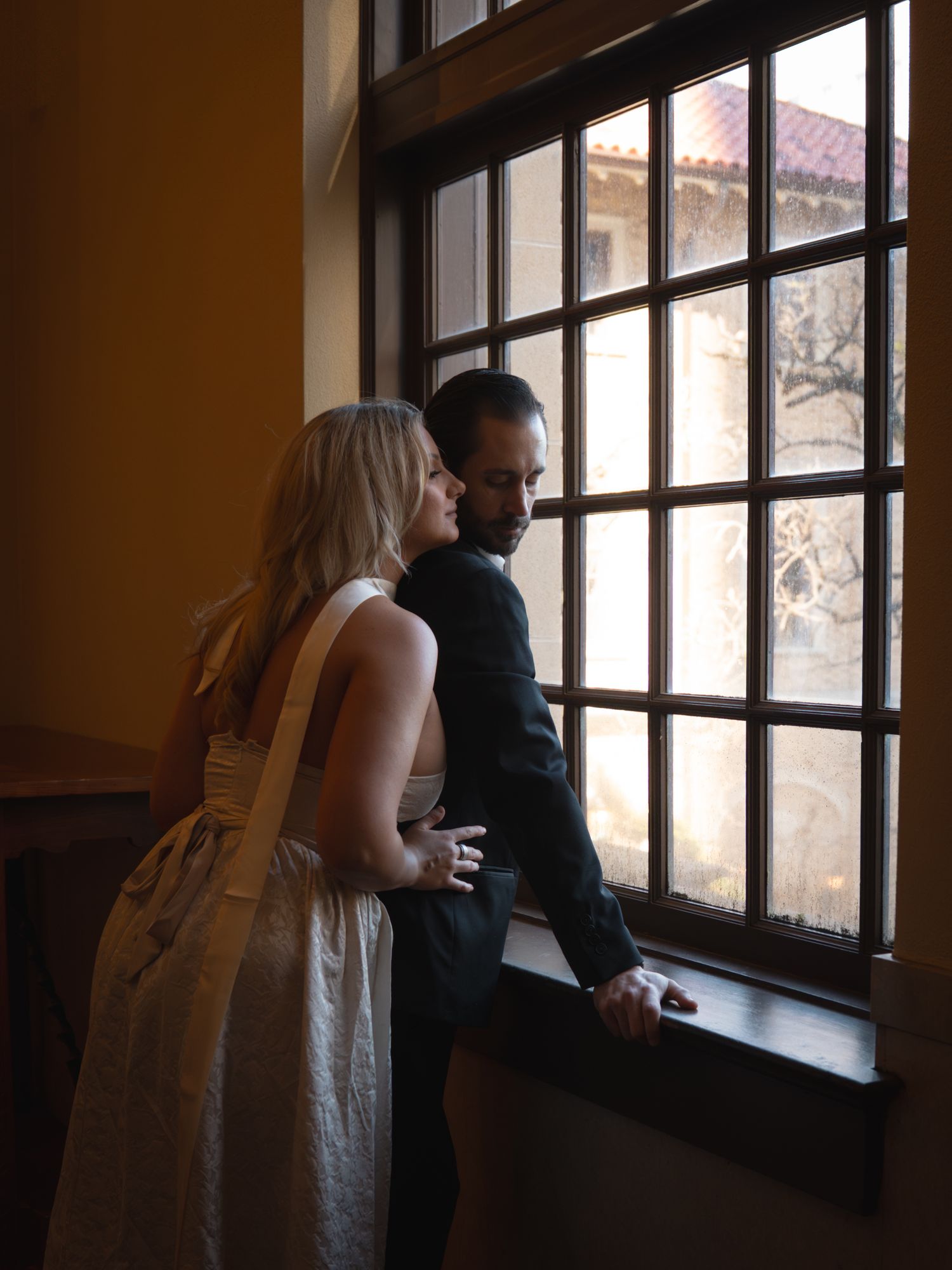 Couple in elegant attire sharing an intimate moment by a large grid window in a warmly lit historic building.