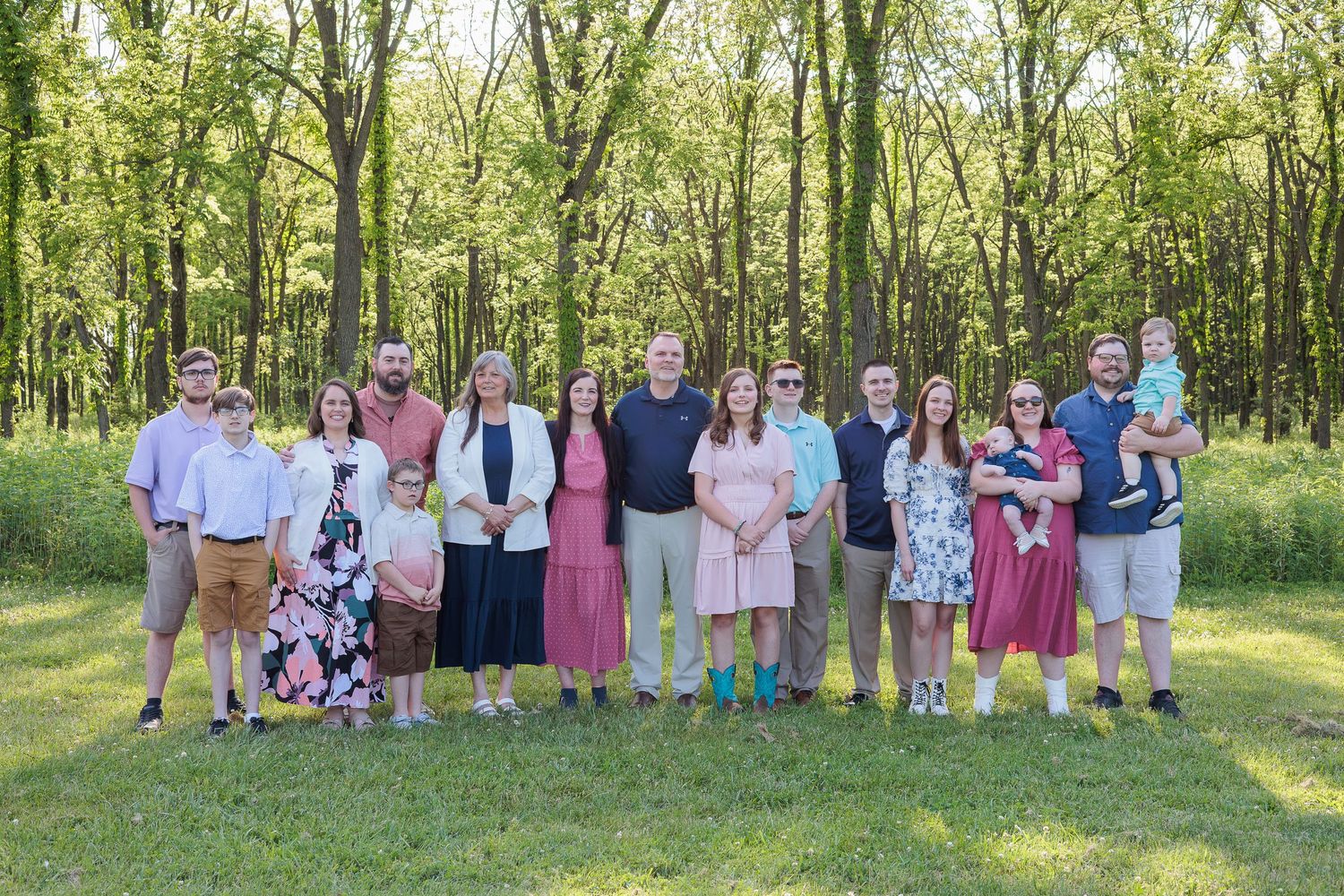 Large extended family group portrait in green wooded setting wearing coordinated spring outfits.