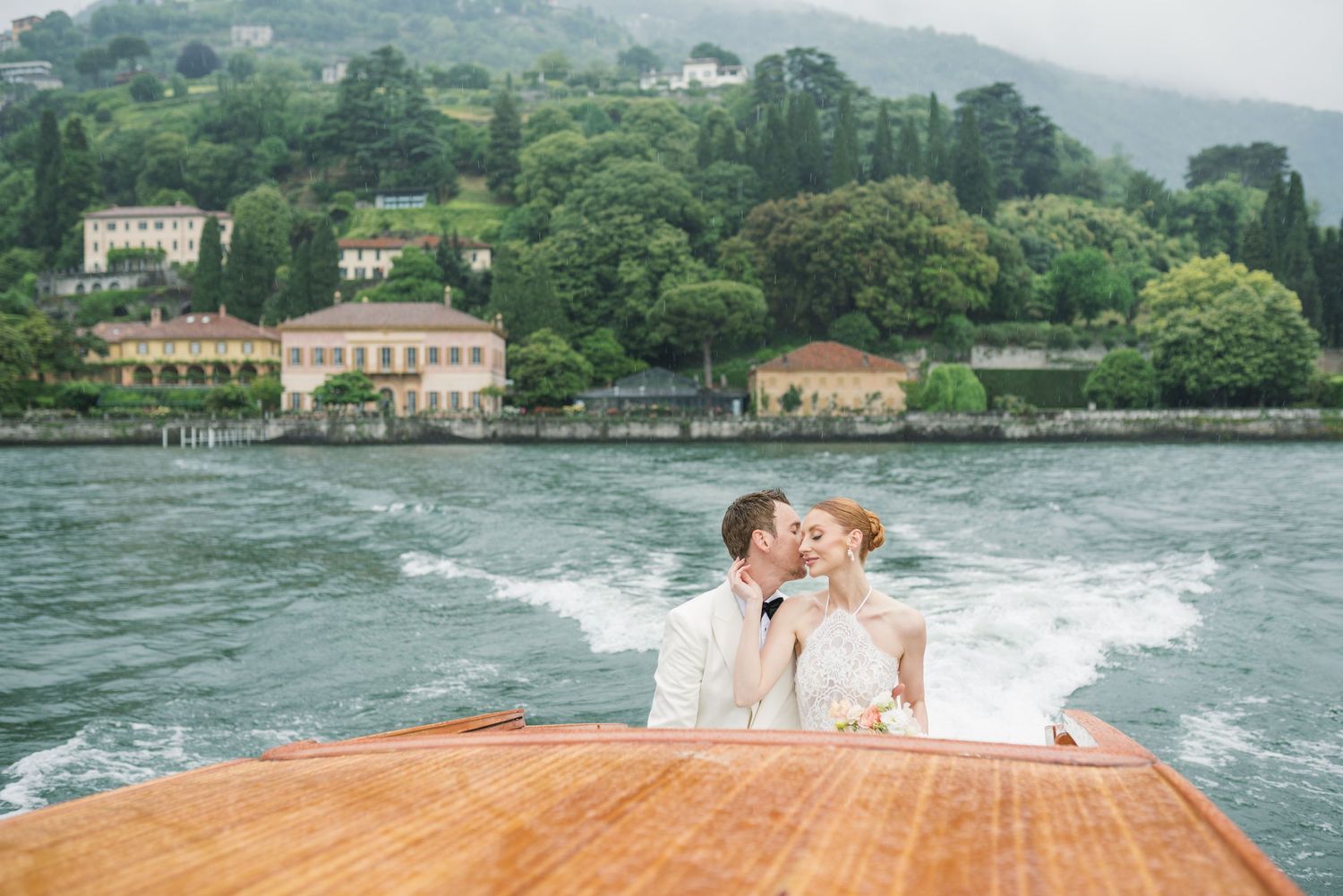 Couple kissing on a wooden speedboat on Lake Como, Italy, with lush green hills and elegant villas in the background.