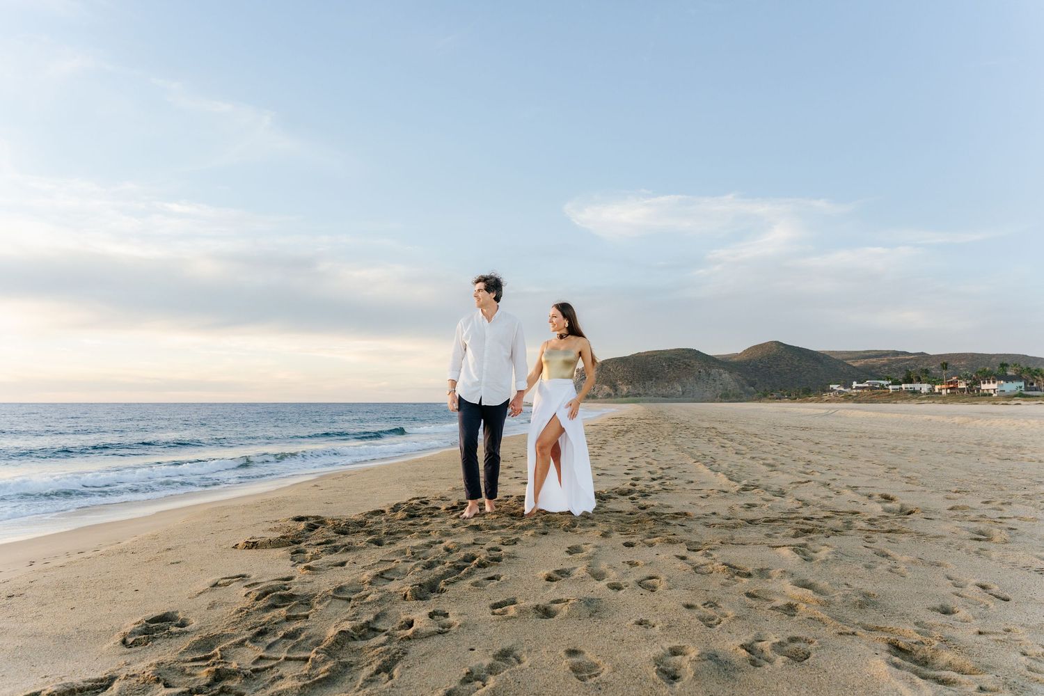 Couple holding hands walking on sandy beach at sunset with ocean waves and hills in background.
