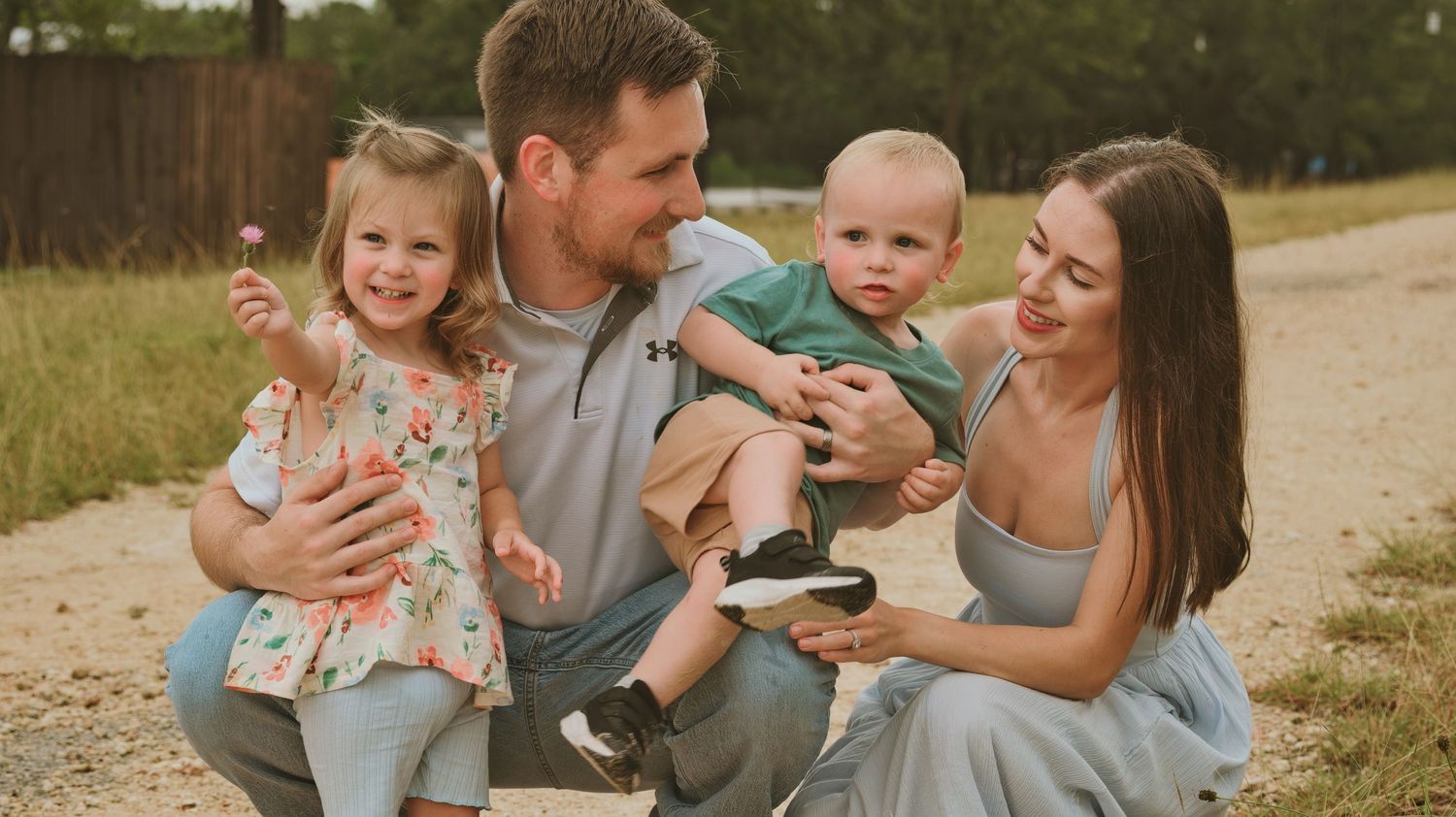 Happy young family with two toddlers smiling outdoors on a dirt path surrounded by greenery.
