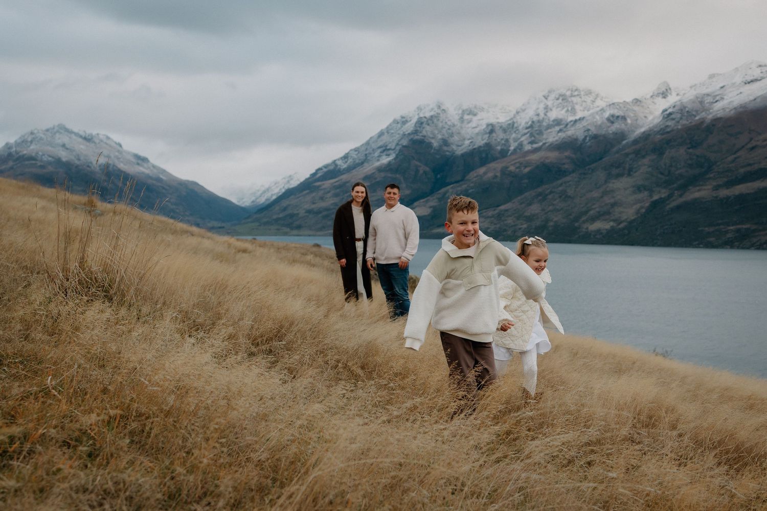 Family of four enjoying a scenic lakeside walk with snow-capped mountains in Queenstown, New Zealand.