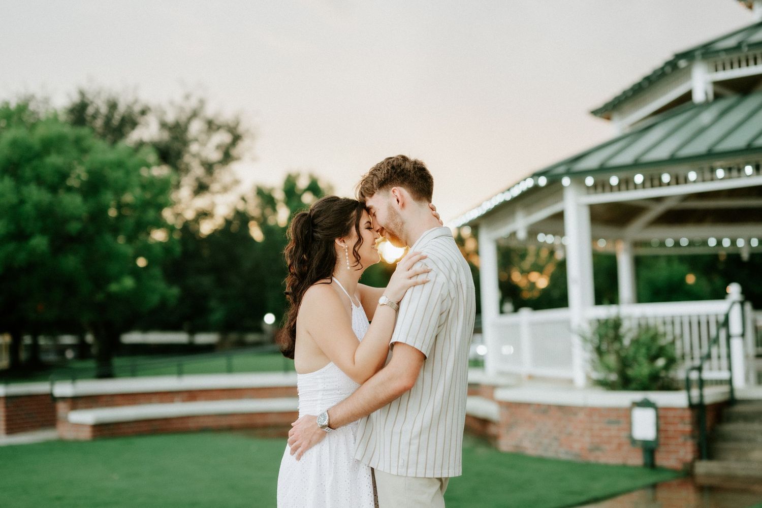 Couple embracing at sunset near a gazebo with string lights, sharing a romantic moment outdoors.