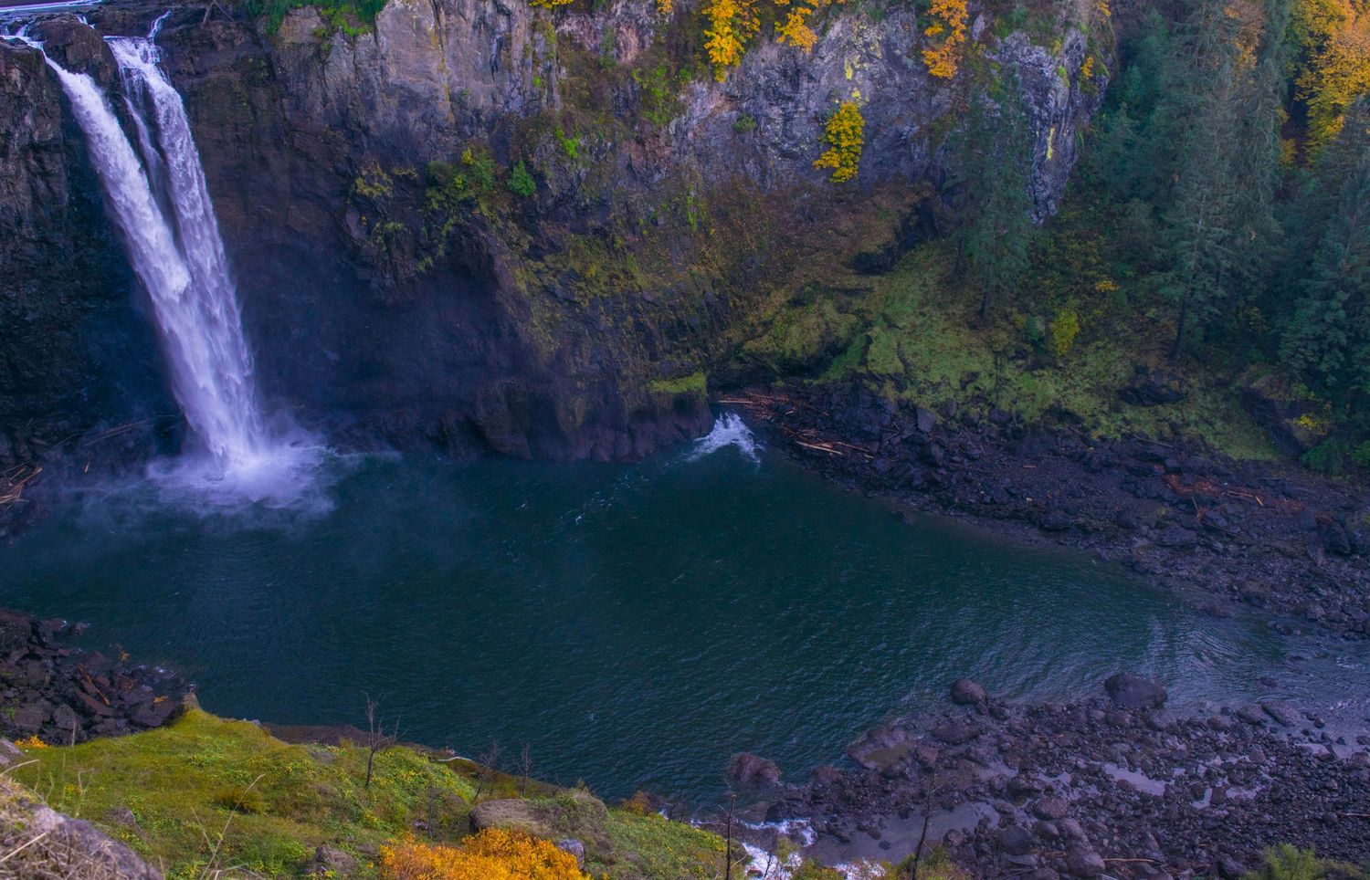 A dramatic waterfall cascades into a deep green pool surrounded by autumn-colored cliffs.