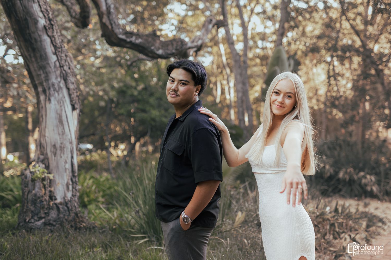 A couple poses playfully back-to-back in a scenic woodland setting.