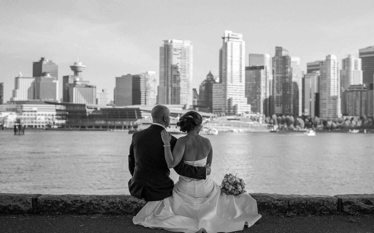 A couple sits together on a waterfront overlooking the Vancouver skyline in black and white photography.