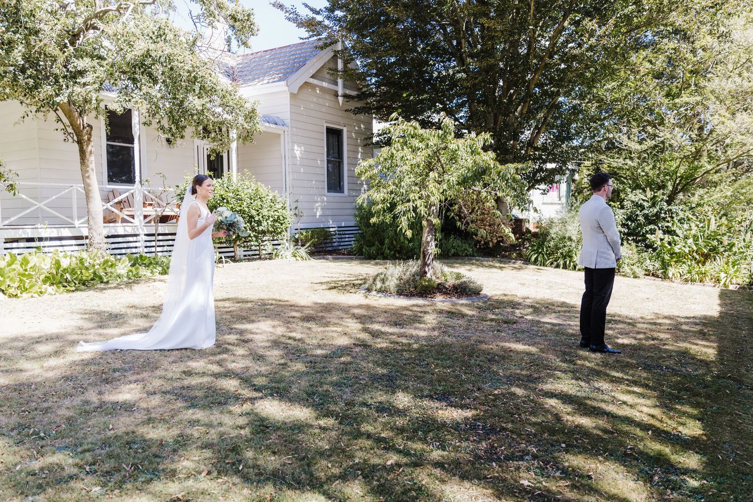 First look wedding photo shoot in front of a white farmhouse with trees.