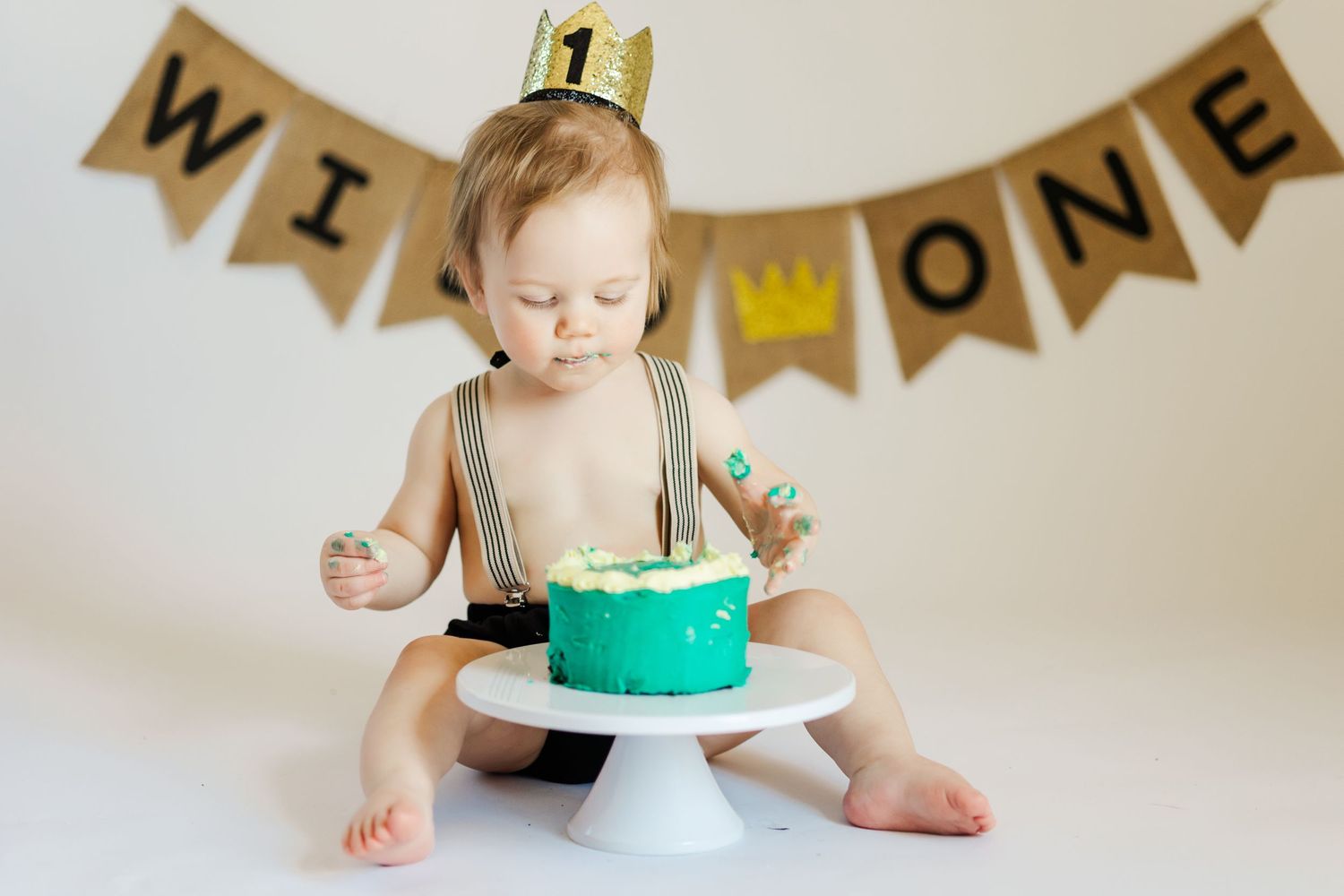 Baby in crown celebrating first birthday with turquoise cake and WIN ONE banner.