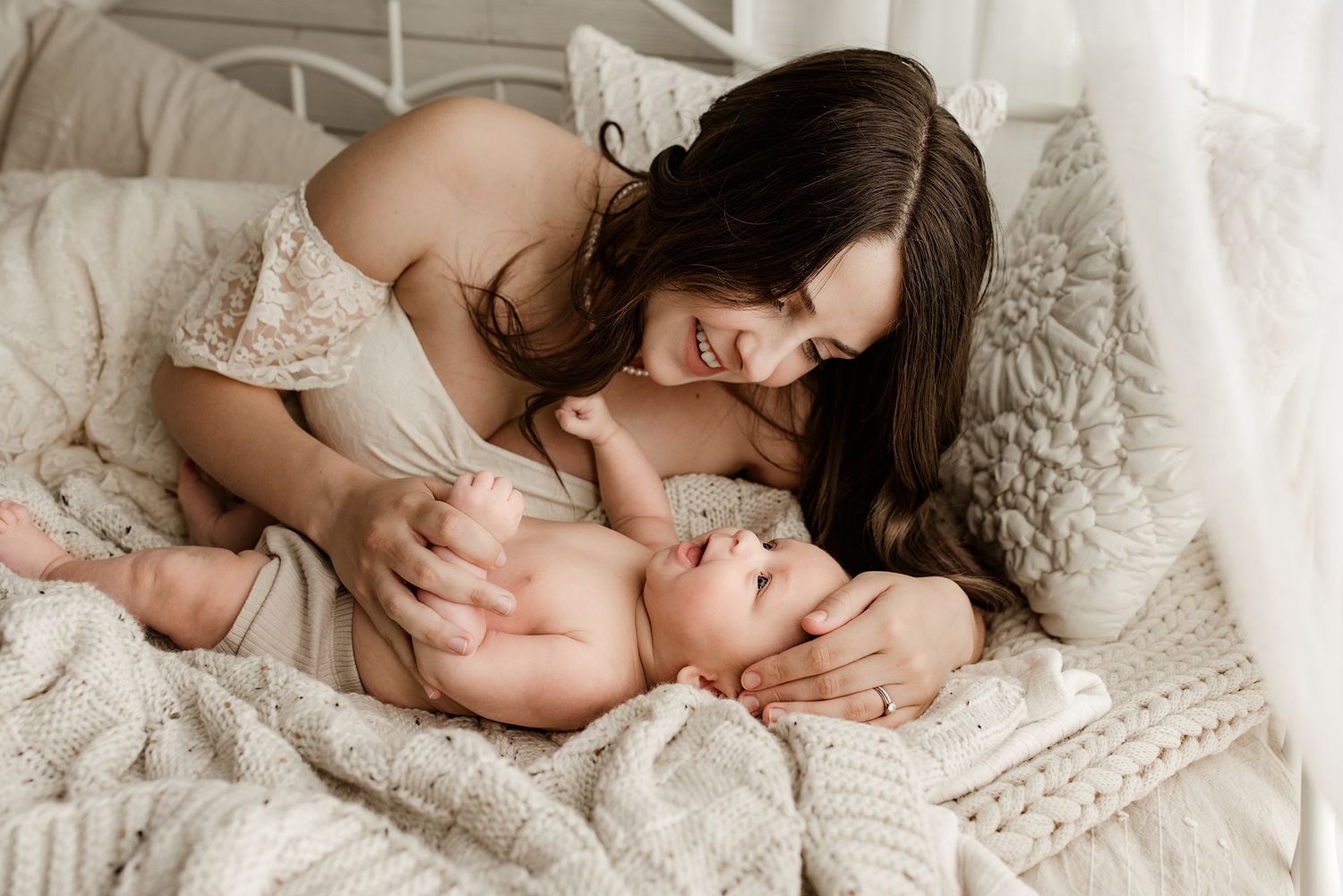 Loving dark-haired mother smiles and plays with happy baby lying on cozy white knit bedding.