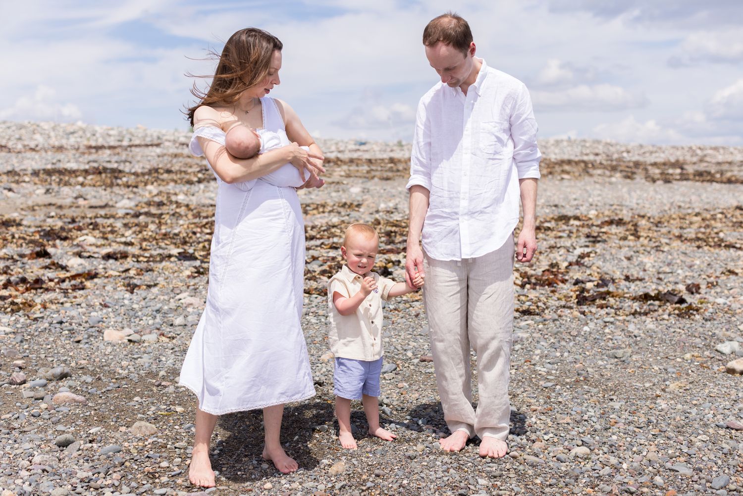 Family of four with a newborn and toddler stand on beach rocks in Winthrop, Massachusetts for family photos on a sunny day.