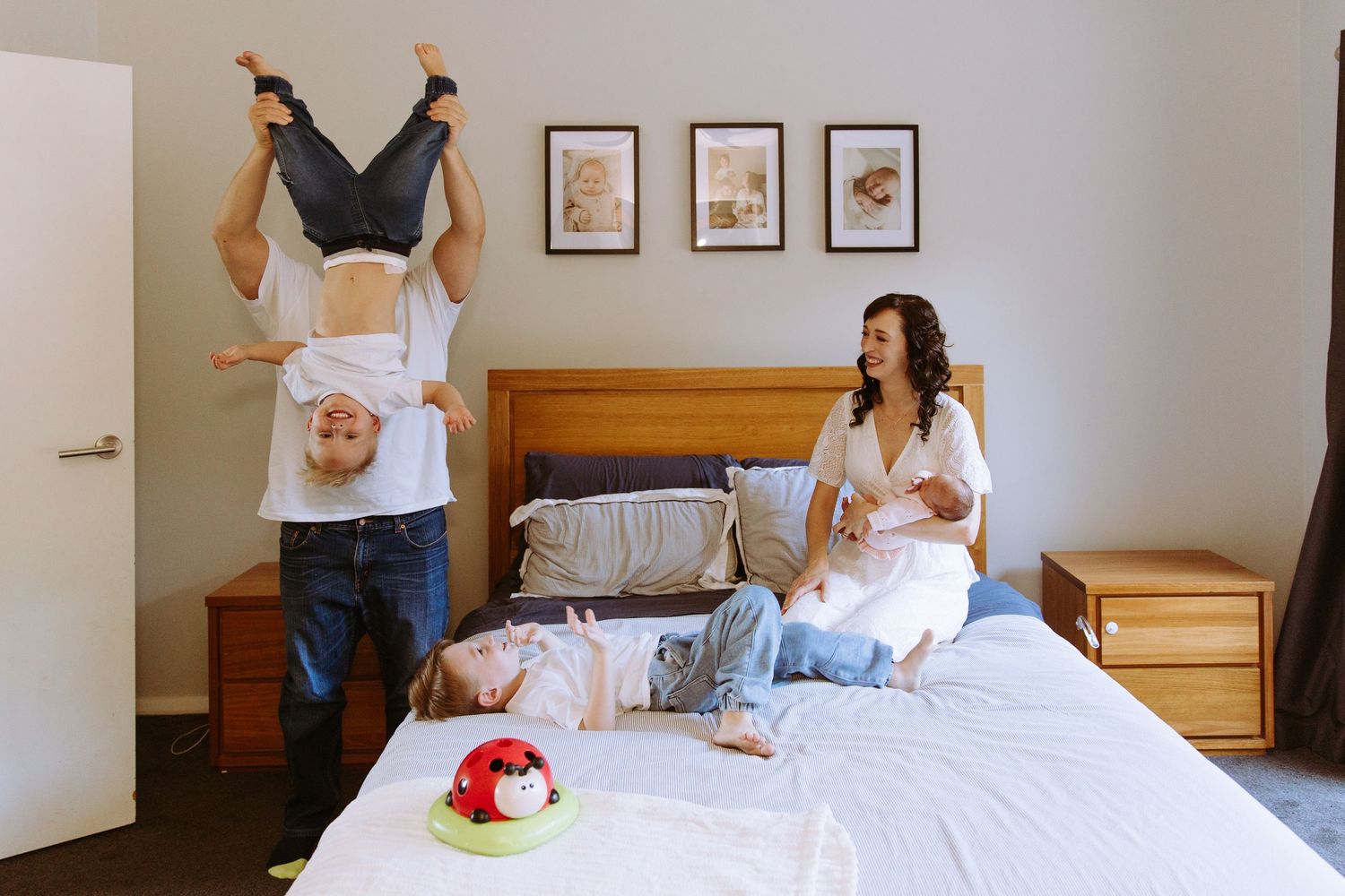 Family playing in bedroom, father holding child upside down while mother sits on bed with baby and toddler.