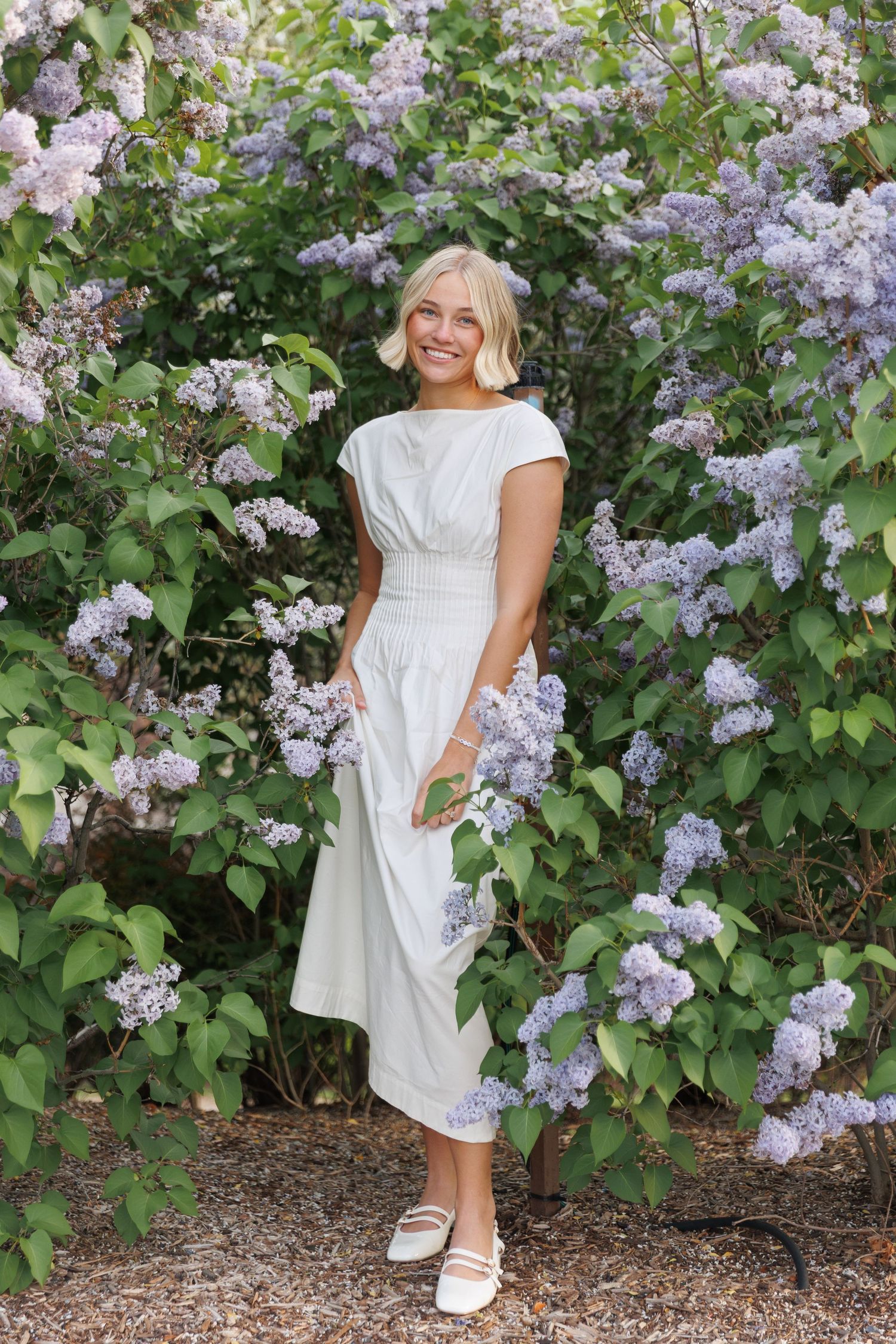 Smiling blonde woman in white midi dress standing among blooming purple lilac bushes.