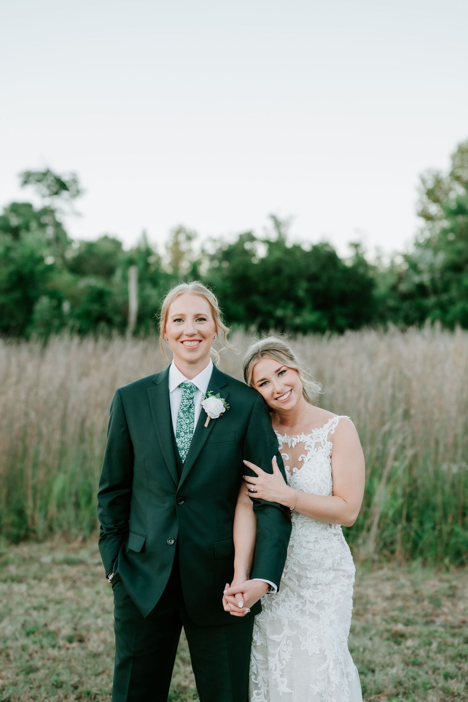 Two smiling brides embrace in a grassy field, one in a dark green suit with boutonniere and one in a white lace wedding gown.