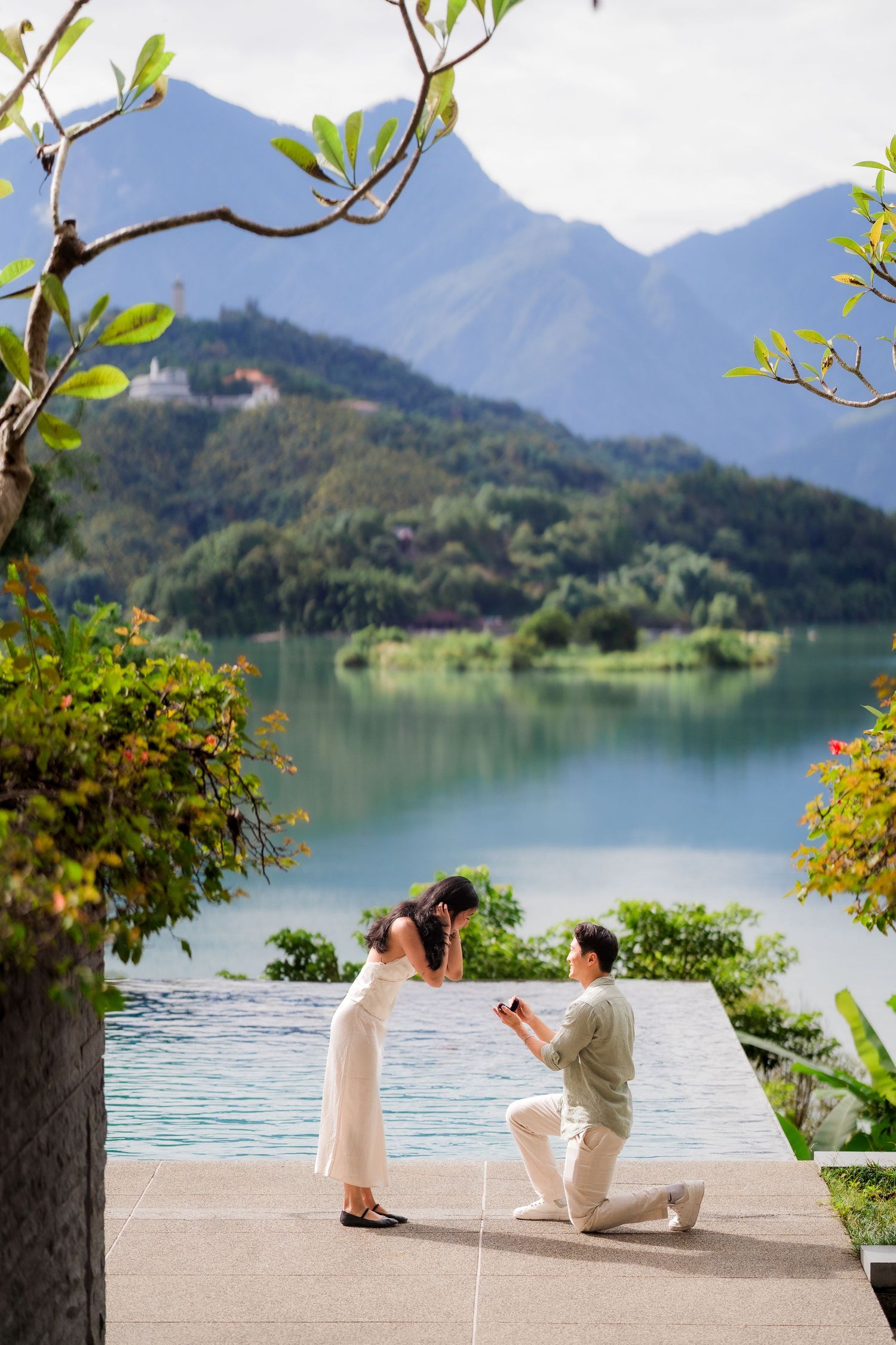 Man proposes to woman on one knee by a serene lake with lush green mountains in the background at The Lalu Hotel at Sun Moon Lake.