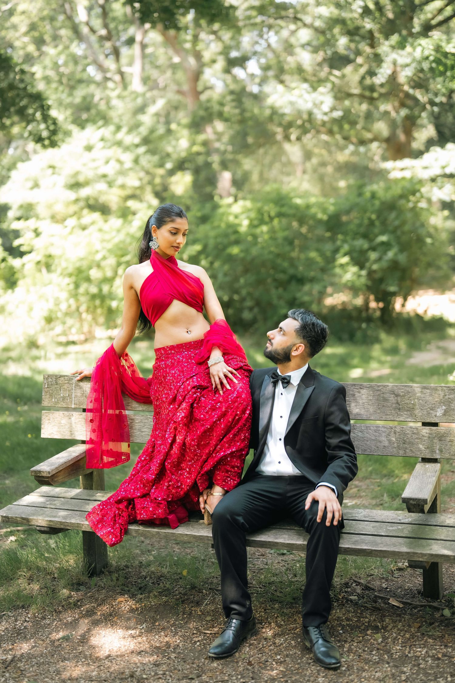Couple in formal attire posing on a park bench, woman in red lehenga and man in black tuxedo. At Richmond Park