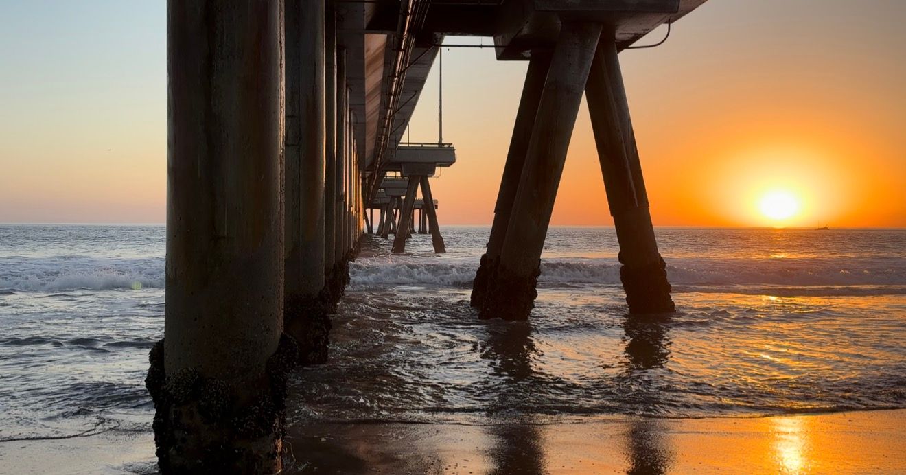 Golden sunset glowing on the horizon beneath a wooden beach pier, with waves rolling onto the wet sand.