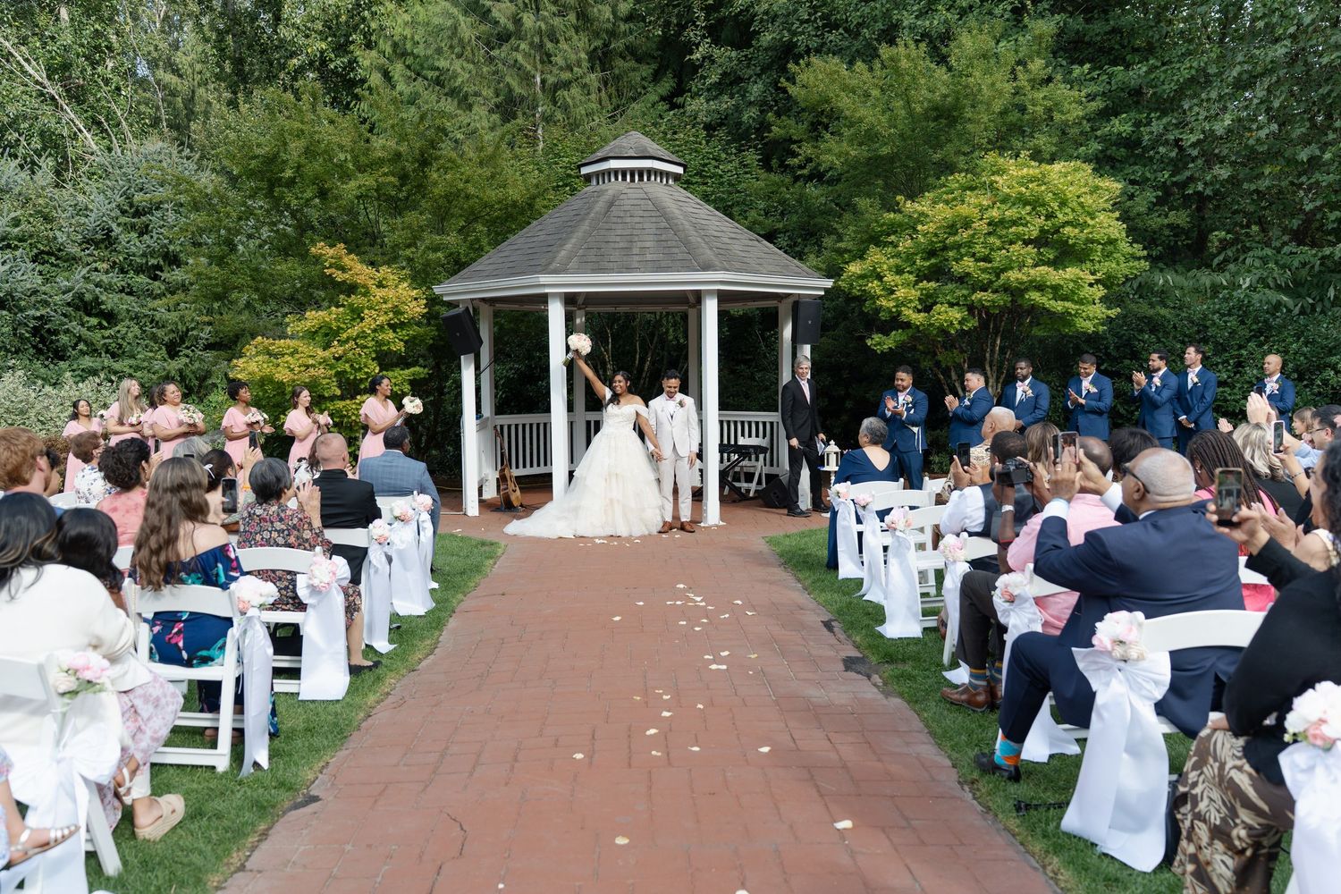 Outdoor wedding ceremony at a gazebo with guests seated on white chairs along a brick aisle surrounded by lush greenery.