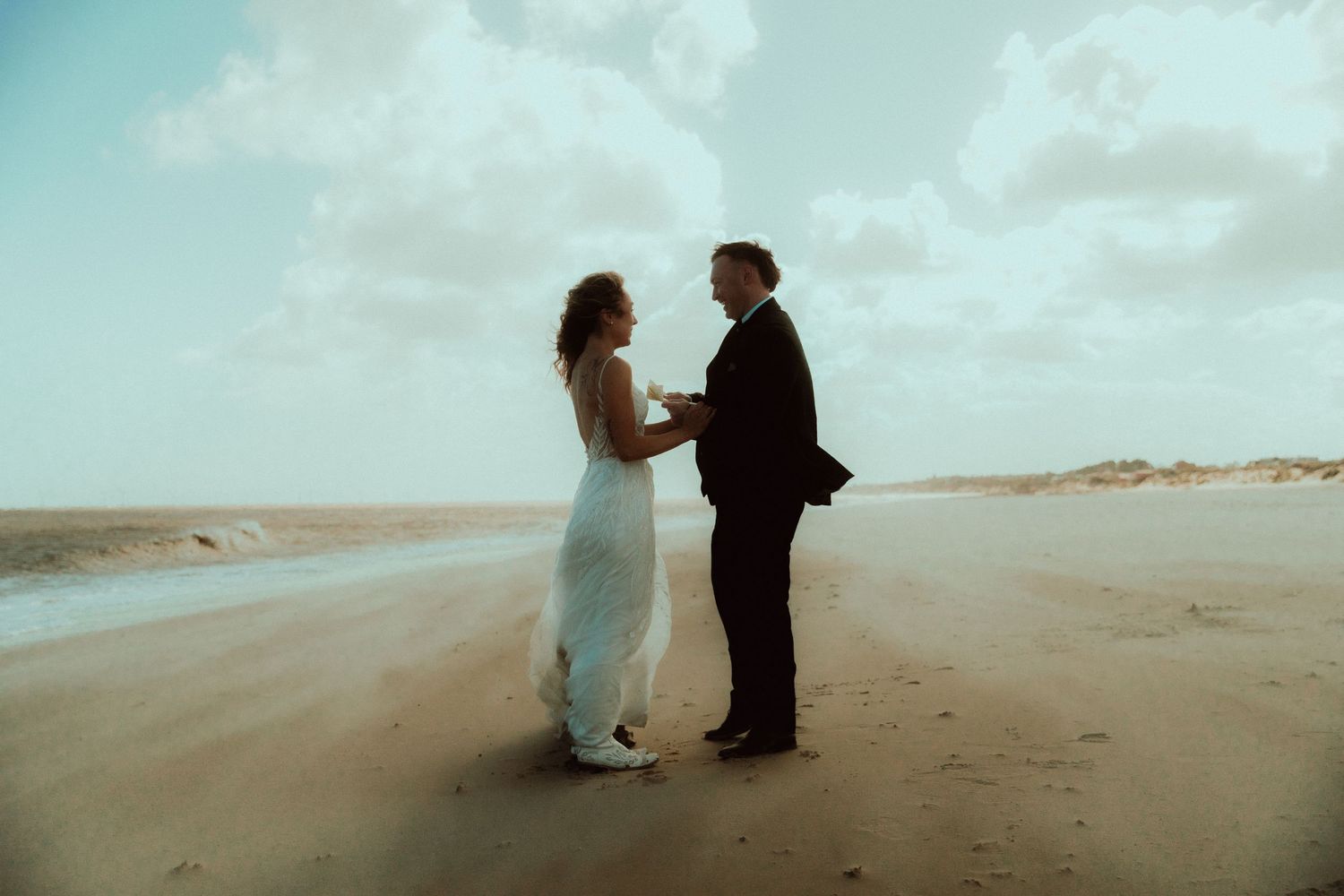 A couple exchanges vows on a sandy Norfolk beach, bride in white gown and groom in black suit, with ocean waves behind them.