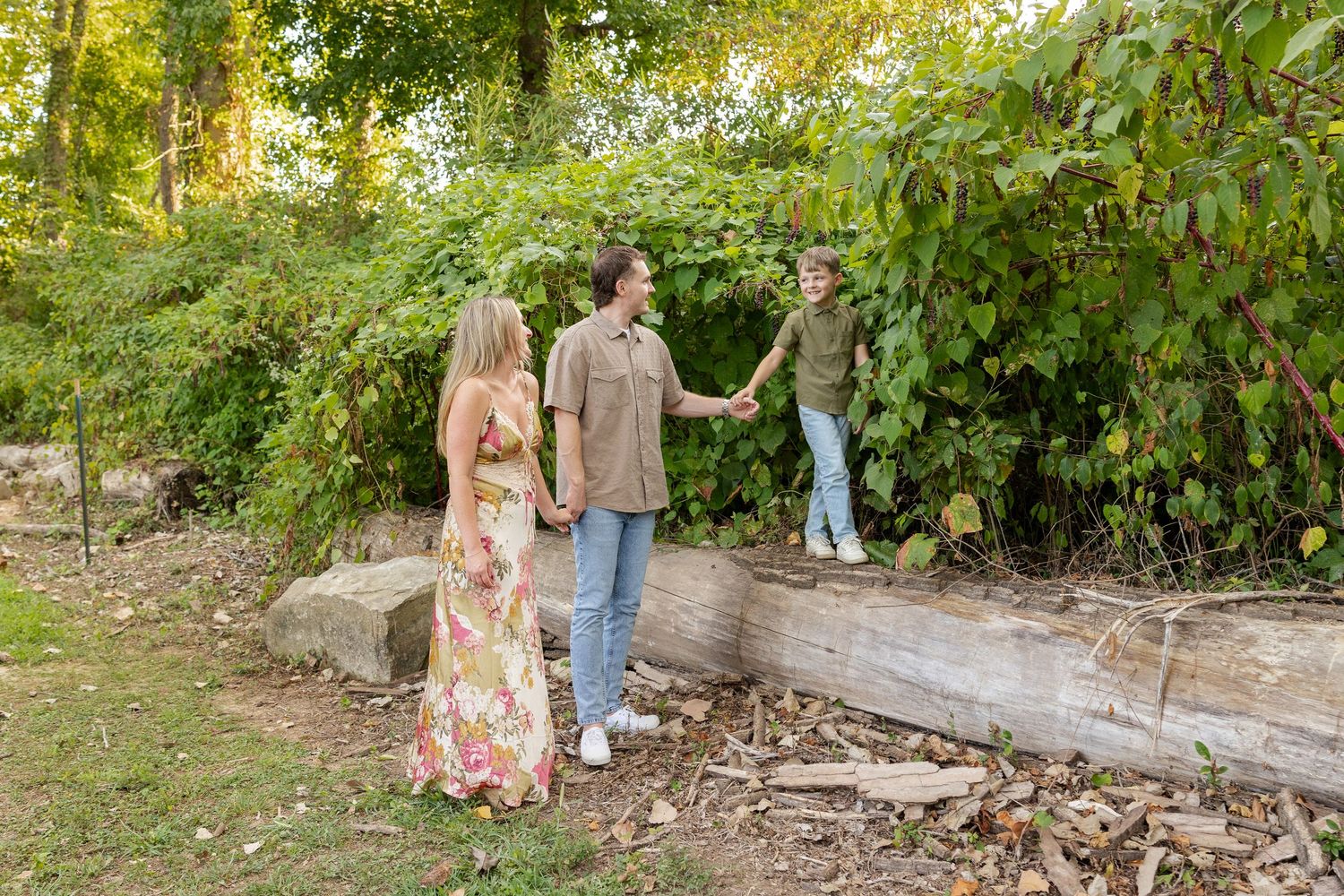 A couple holds hands with a friend standing on a log in a lush green outdoor setting.