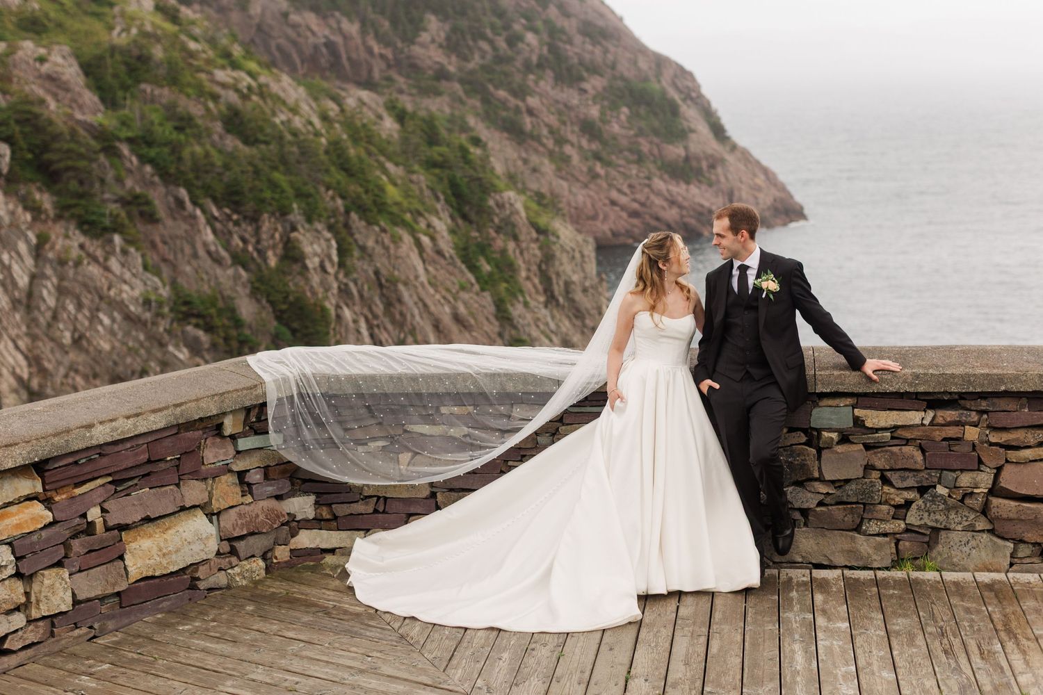 Bride and groom share a romantic moment on a coastal stone terrace, her veil flowing in the wind with ocean views behind.