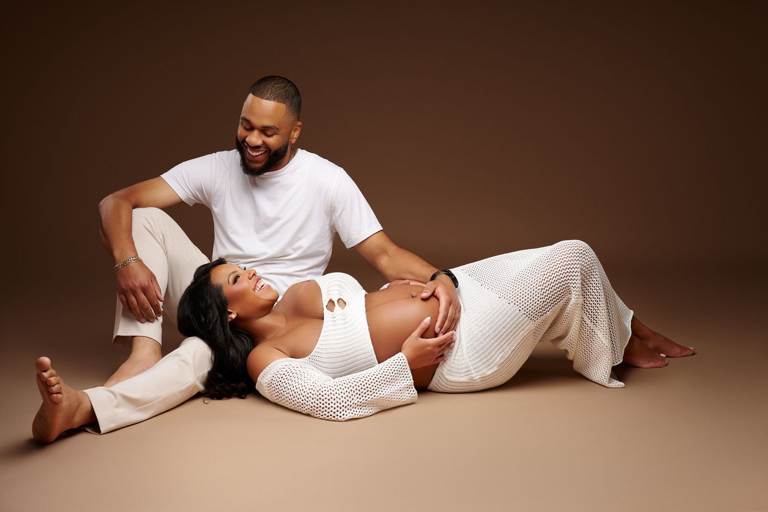 A couple in white outfits poses on a brown backdrop, the pregnant woman reclining as her partner smiles at her.