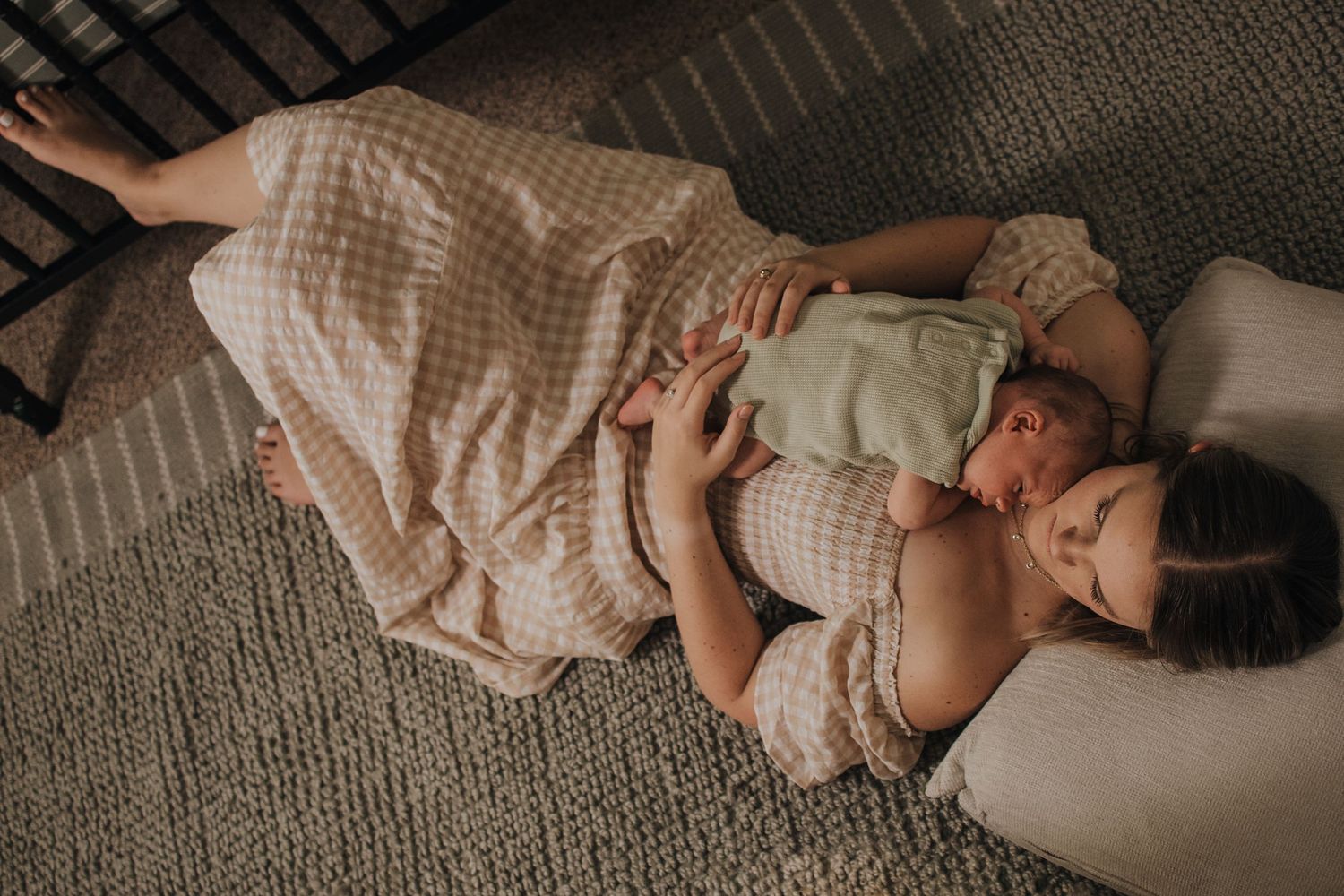 Mother lying on floor cradling newborn baby wrapped in green blanket on checkered bedding.