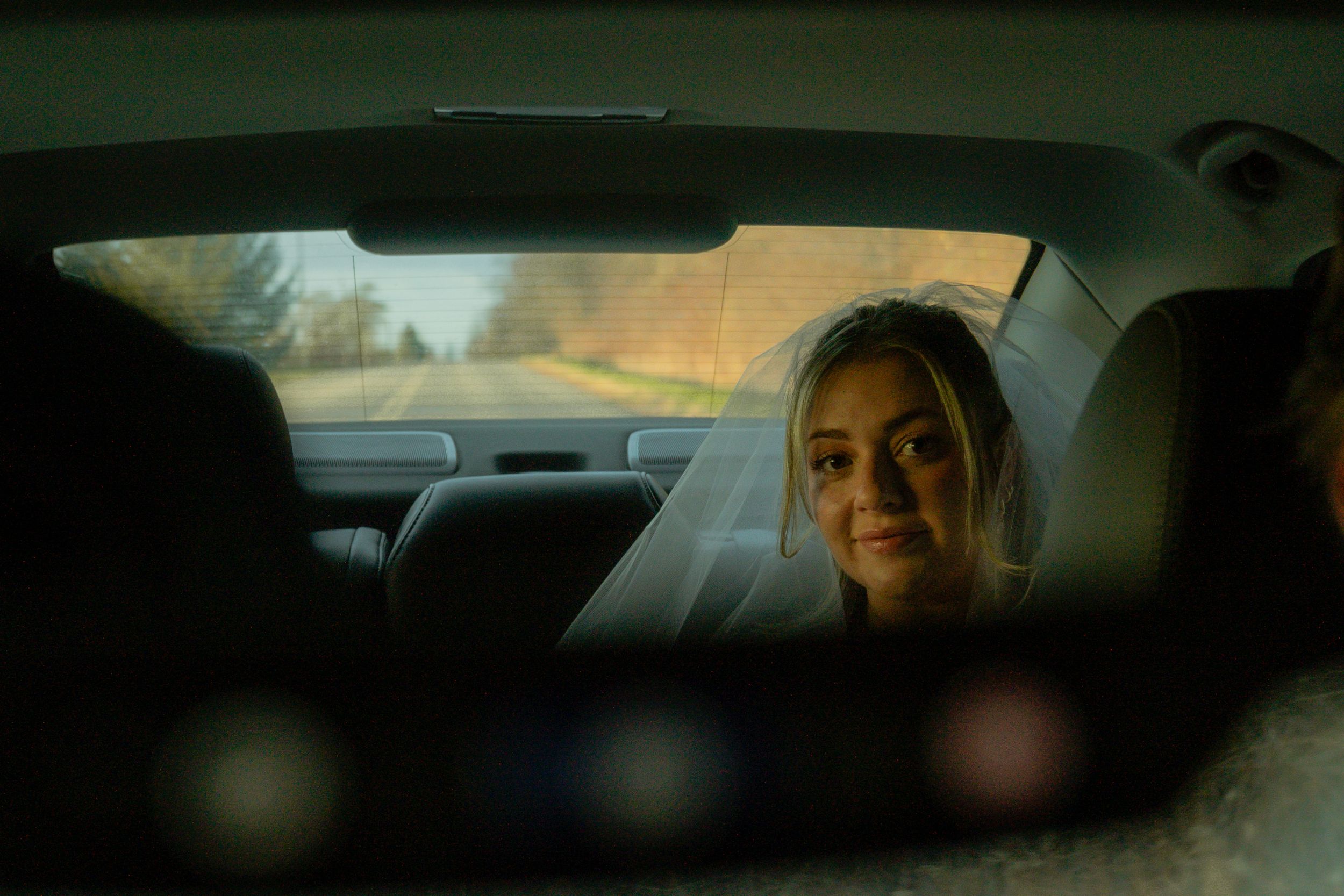 View through a car rearview mirror of a passenger in the backseat smiling at dusk.