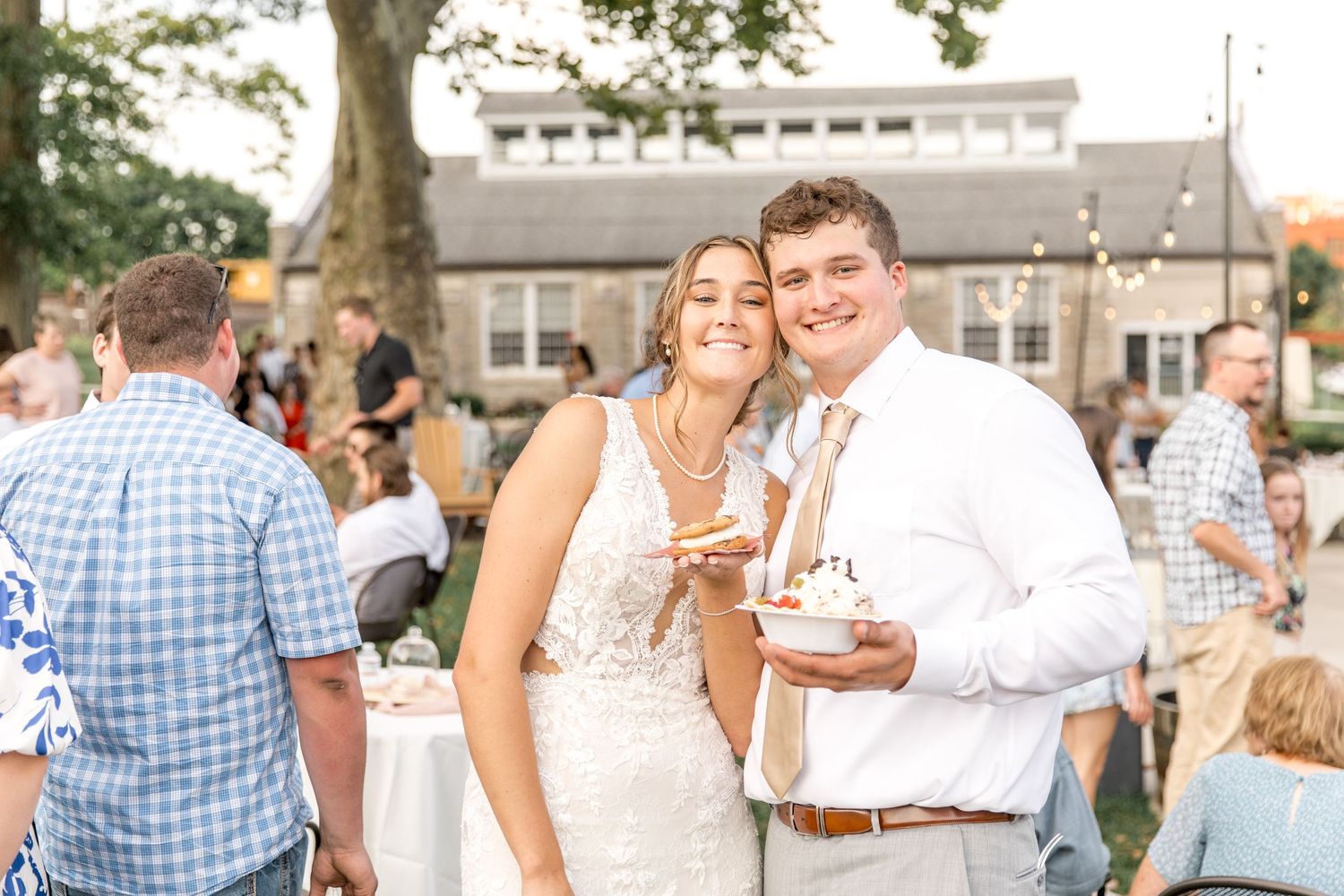 A couple celebrates at an outdoor wedding reception with champagne glasses while guests mingle in the background.