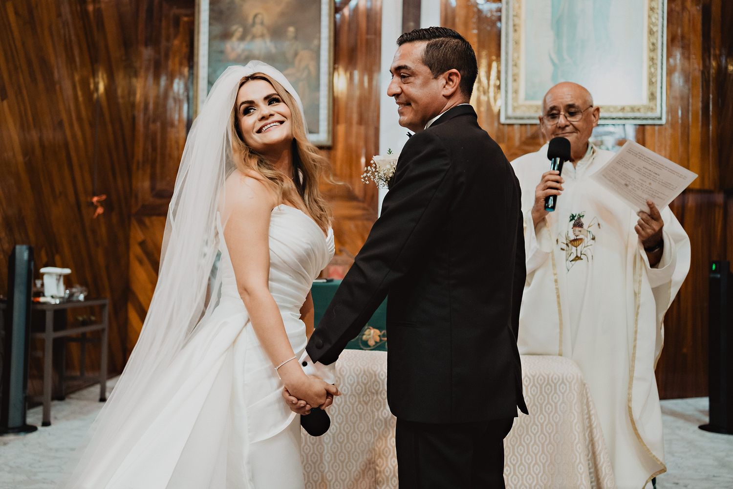 Novios tomados de la mano, novia sonriendo con su padre de fondo