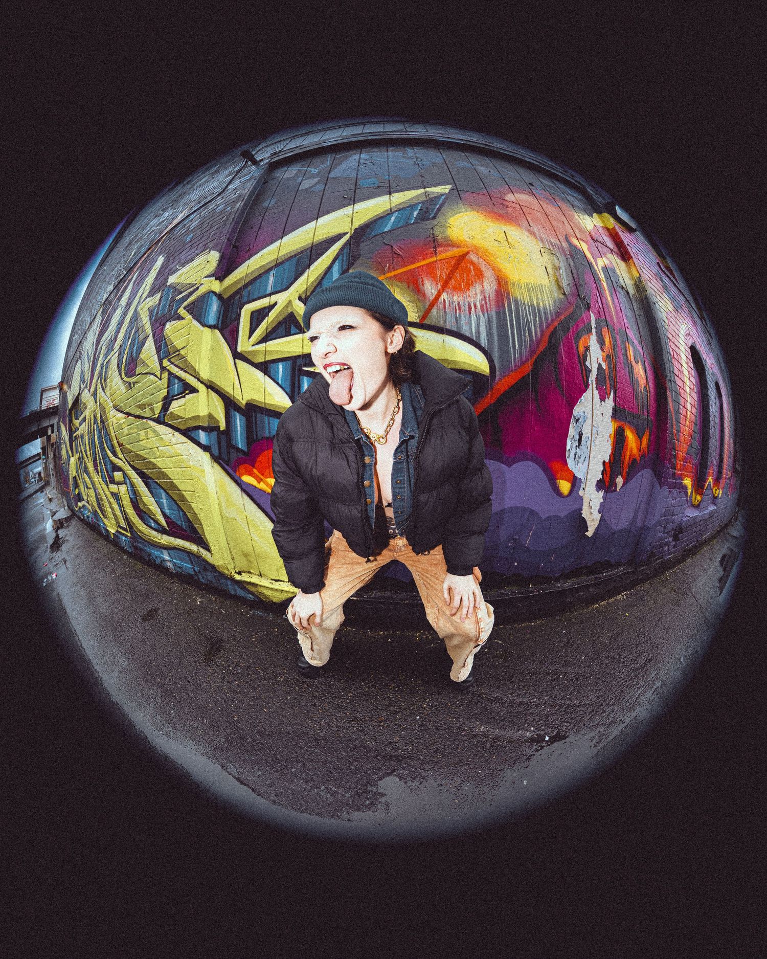 A fisheye lens photo shows someone crouching in front of vibrant graffiti art featuring yellow and red designs on a dark wall.