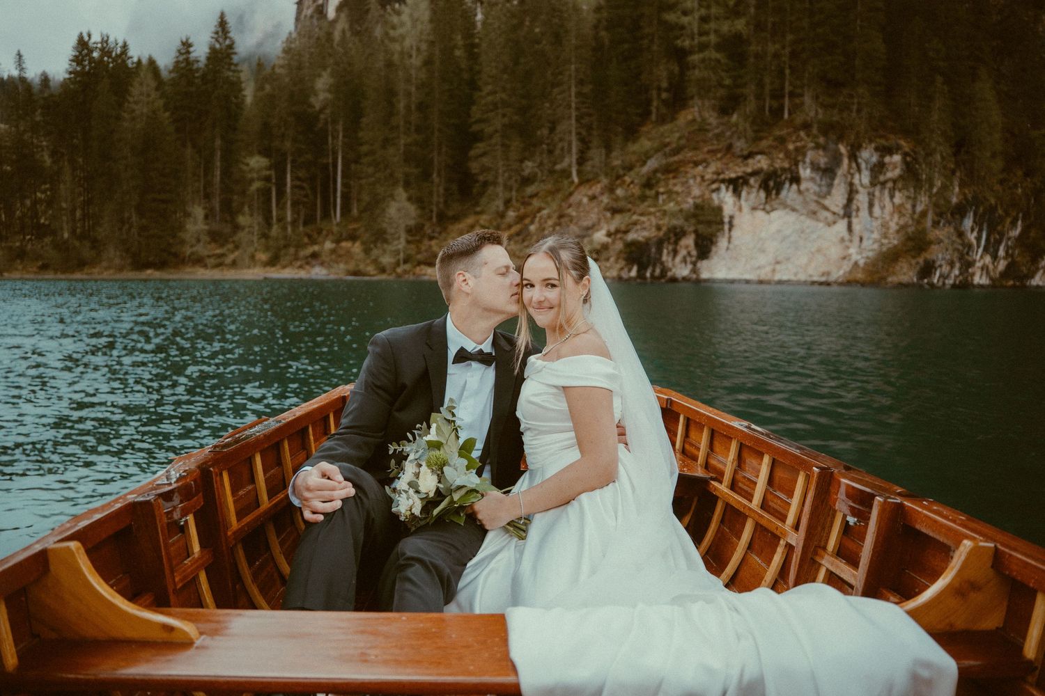 Couple in wedding attire sharing romantic moments in wooden canoe on serene mountain lake during sunset.
