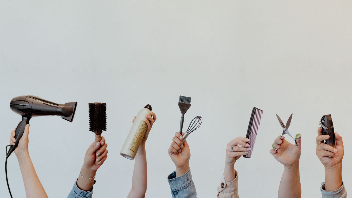 Multiple hands holding up various hairstyling tools and products against white background.
