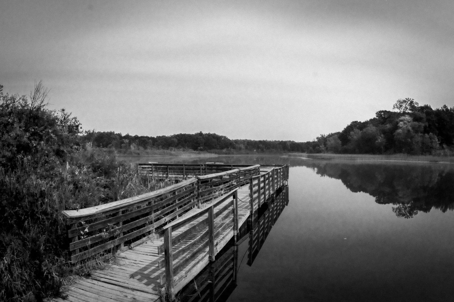 A wooden boardwalk extends over still waters reflecting the cloudy sky in black and white.