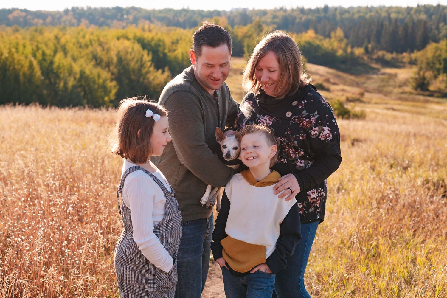 Family shares joyful moment together in golden autumn field with mountains and forest in background.