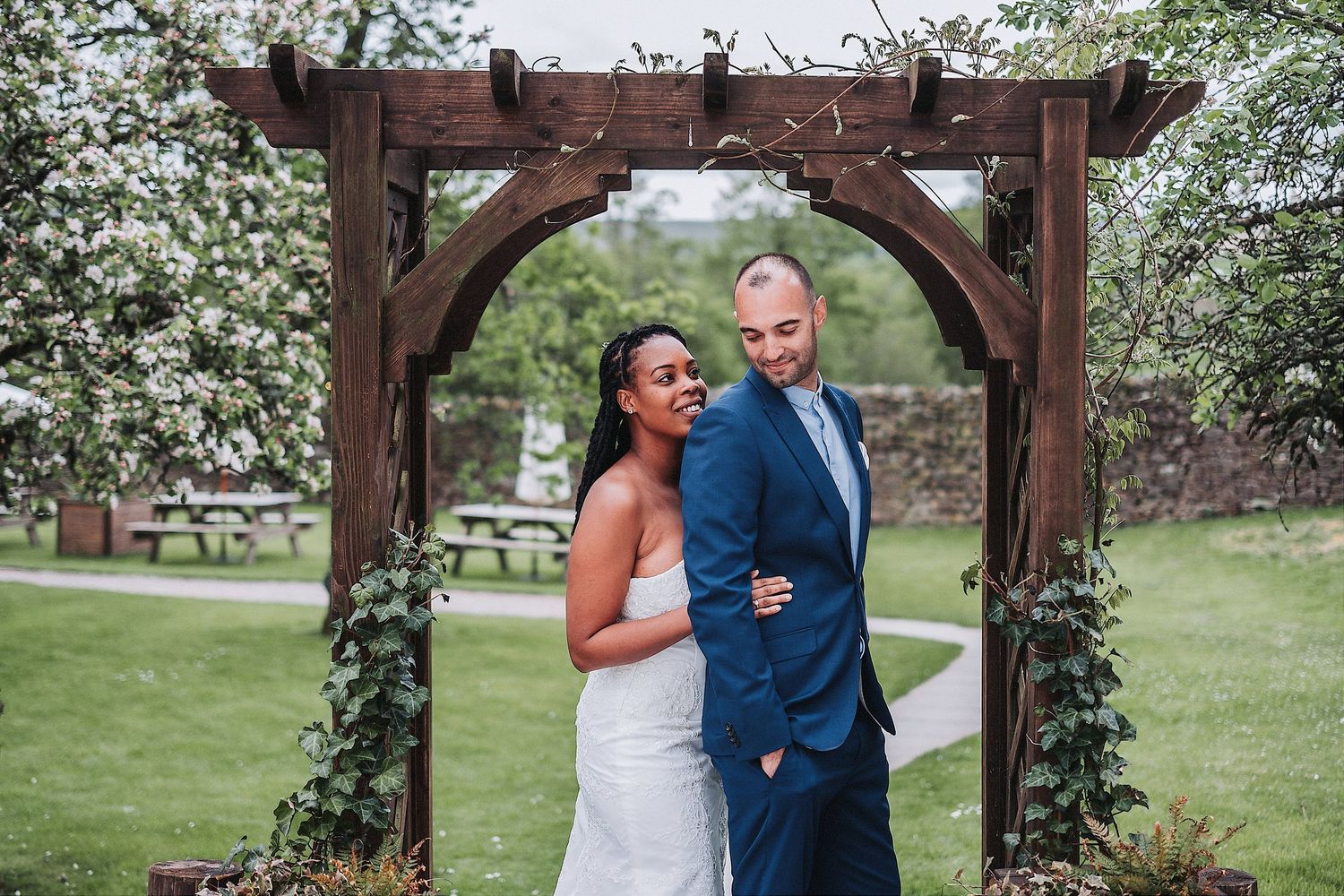 A newlywed couple walks hand in hand down a dirt path bordered by rustic wooden fencing and trees.