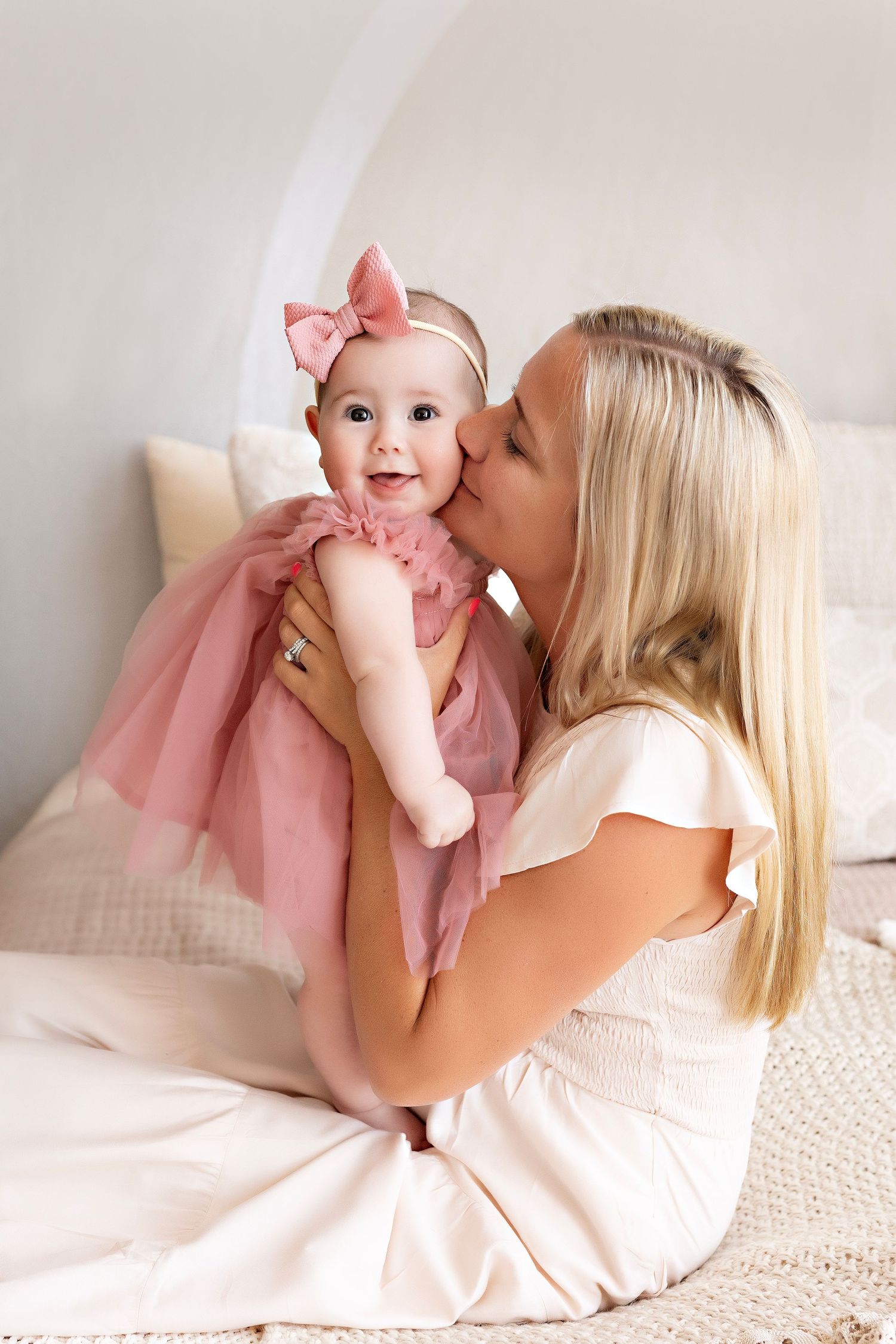 A woman in a white dress shares a tender moment with a baby wearing a pink tulle dress and bow on a light backdrop.