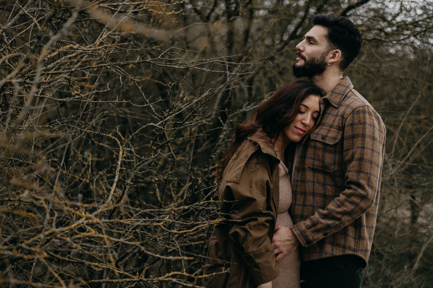 A couple in casual clothing embraces among bare winter branches and dried foliage.