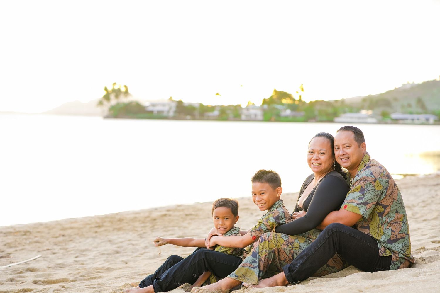 Family sitting together and enjoying a peaceful sunset moment on a sandy beach beside calm waters and hills.