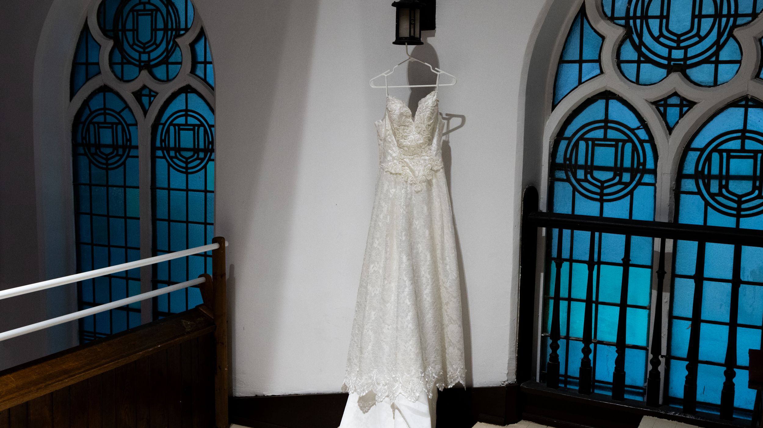 A white lace wedding dress hangs in front of blue stained glass church windows creating an ethereal silhouette.