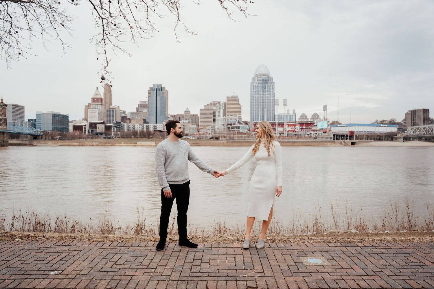 A couple holds hands by the Ohio River with the Cincinnati skyline visible across the water on a cloudy day.