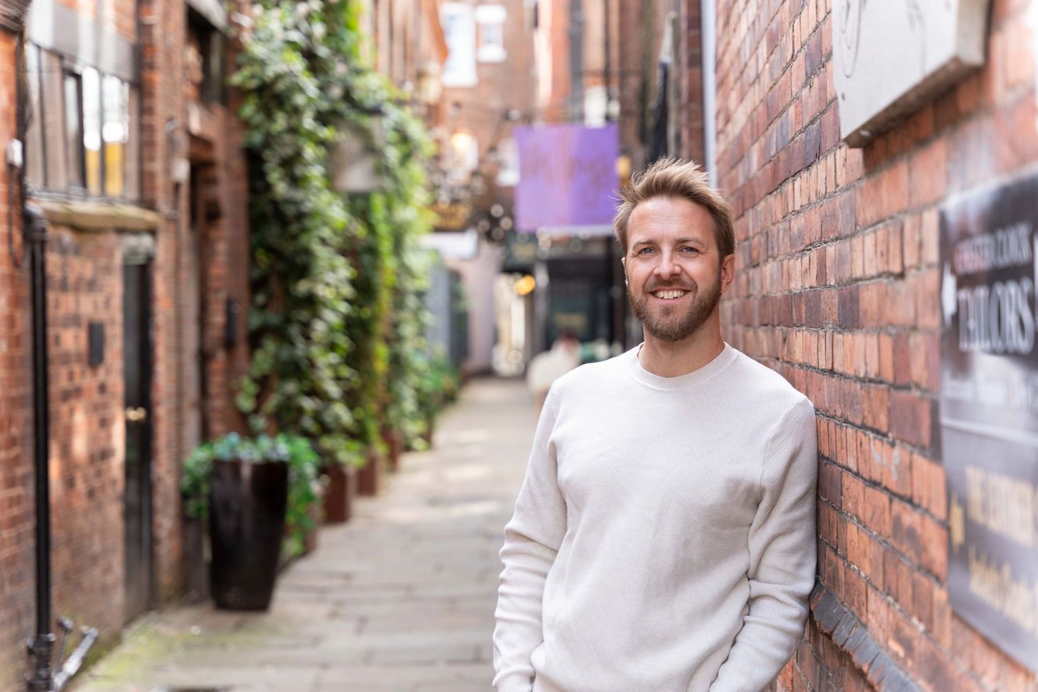 A male entrepreneur in a white sweater smiles against a brick all on a personal branding photoshoot with Steve Currie Photography