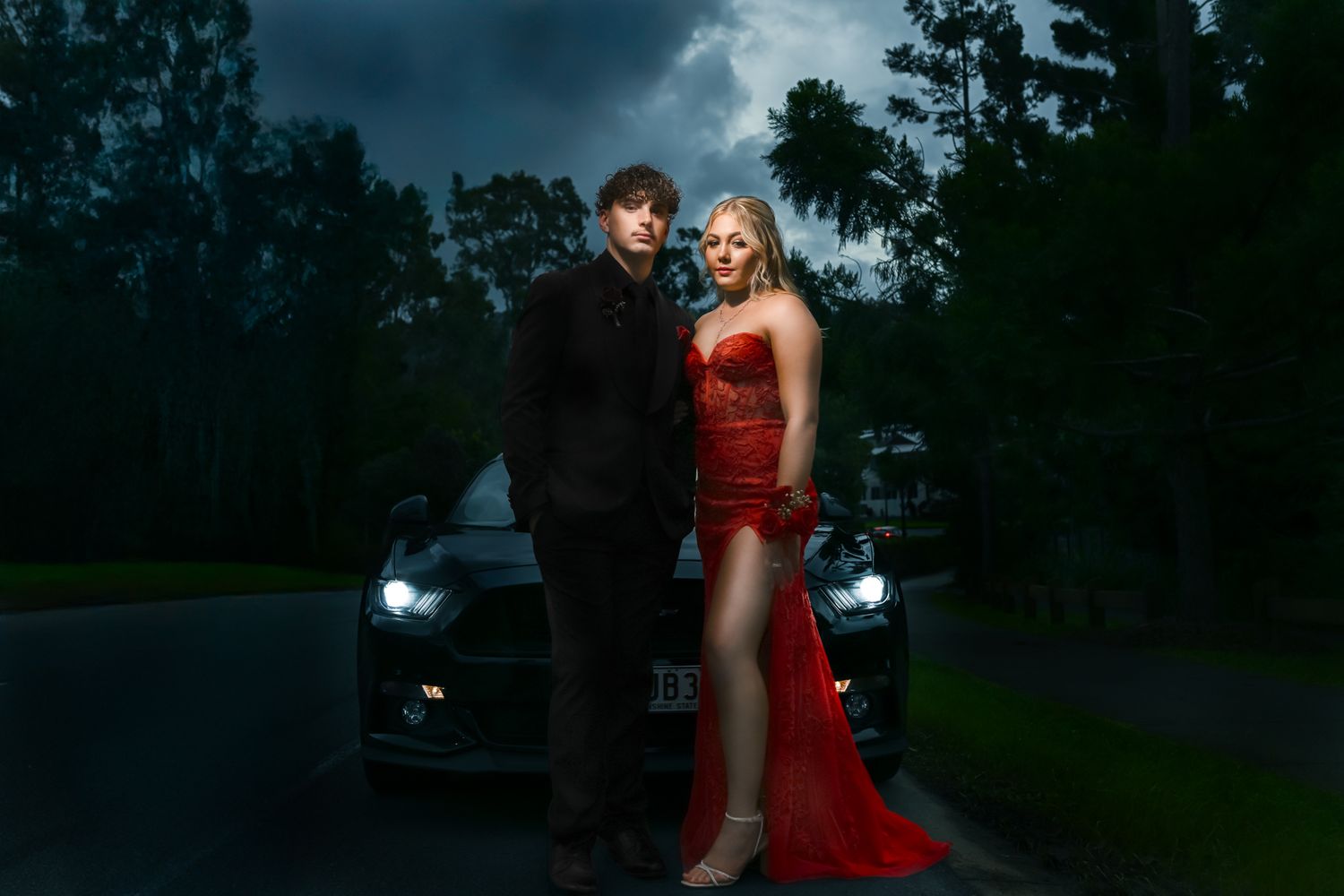 A couple in formal attire poses in front of a dark car at dusk with moody lighting and trees in the background.