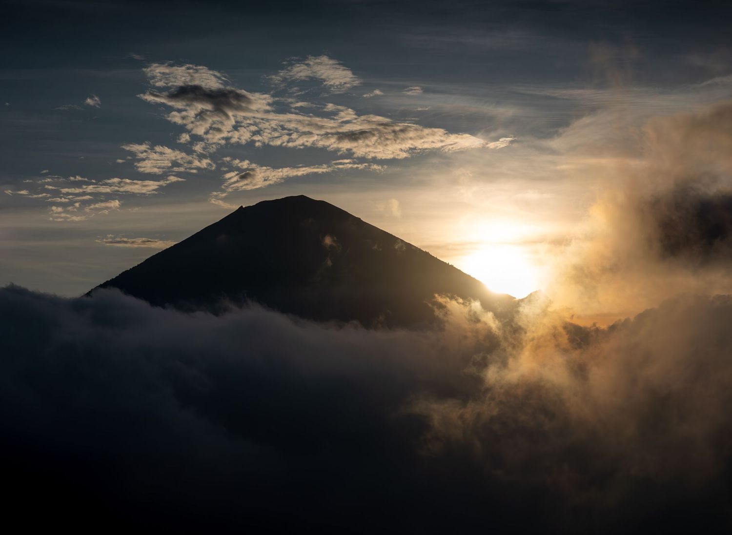 Dark silhouette of a mountain peak with dramatic sunset and clouds creating a moody atmosphere.